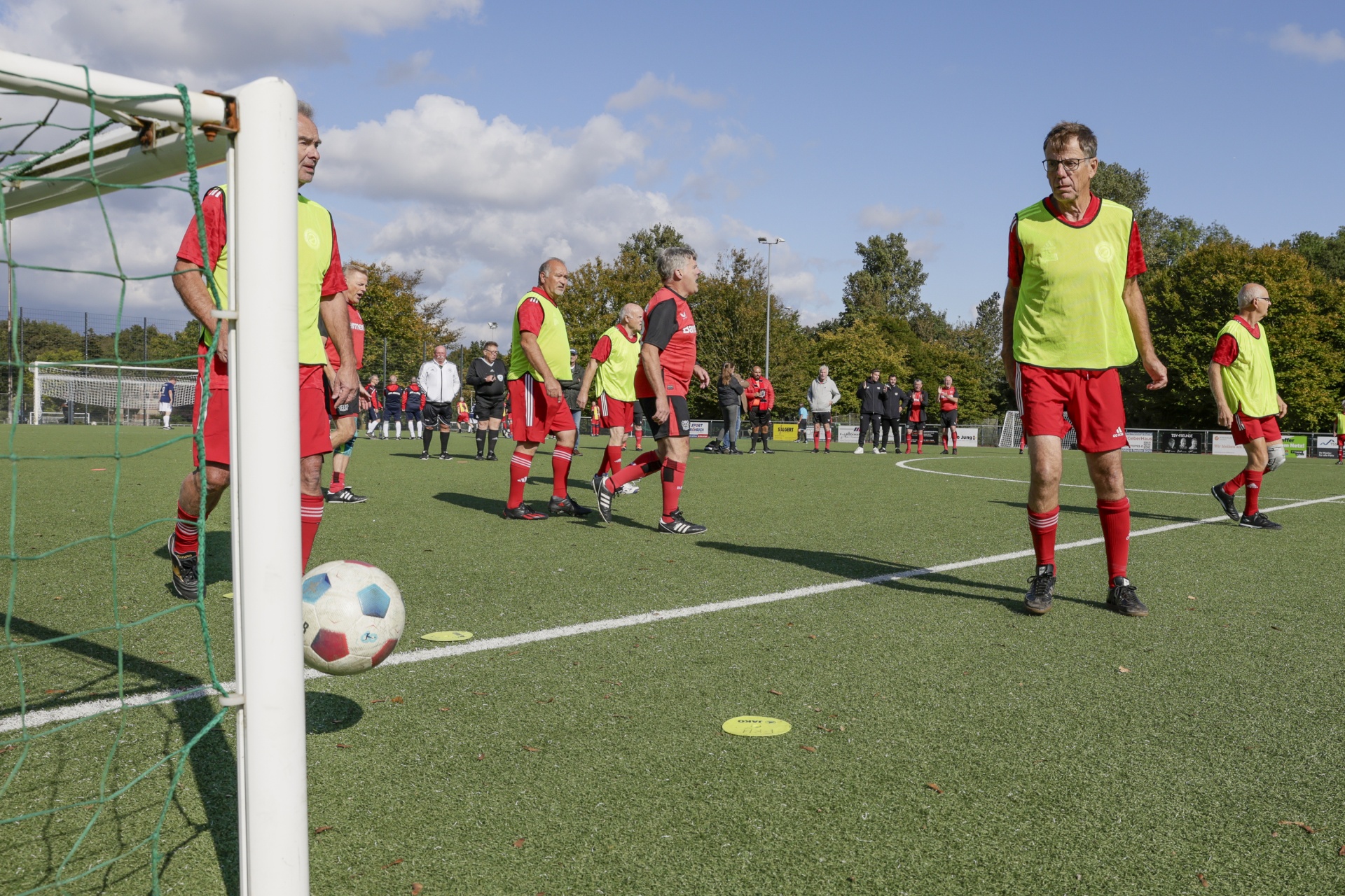 [{"clang_id":1,"value":"Fußballfeld mit Spielern in roten Trikots und gelben Westen. Ein Spieler bereitet einen Schuss vor, der Ball liegt vor dem Tor. Hintergrund: weitere Spieler und Bäume."}]