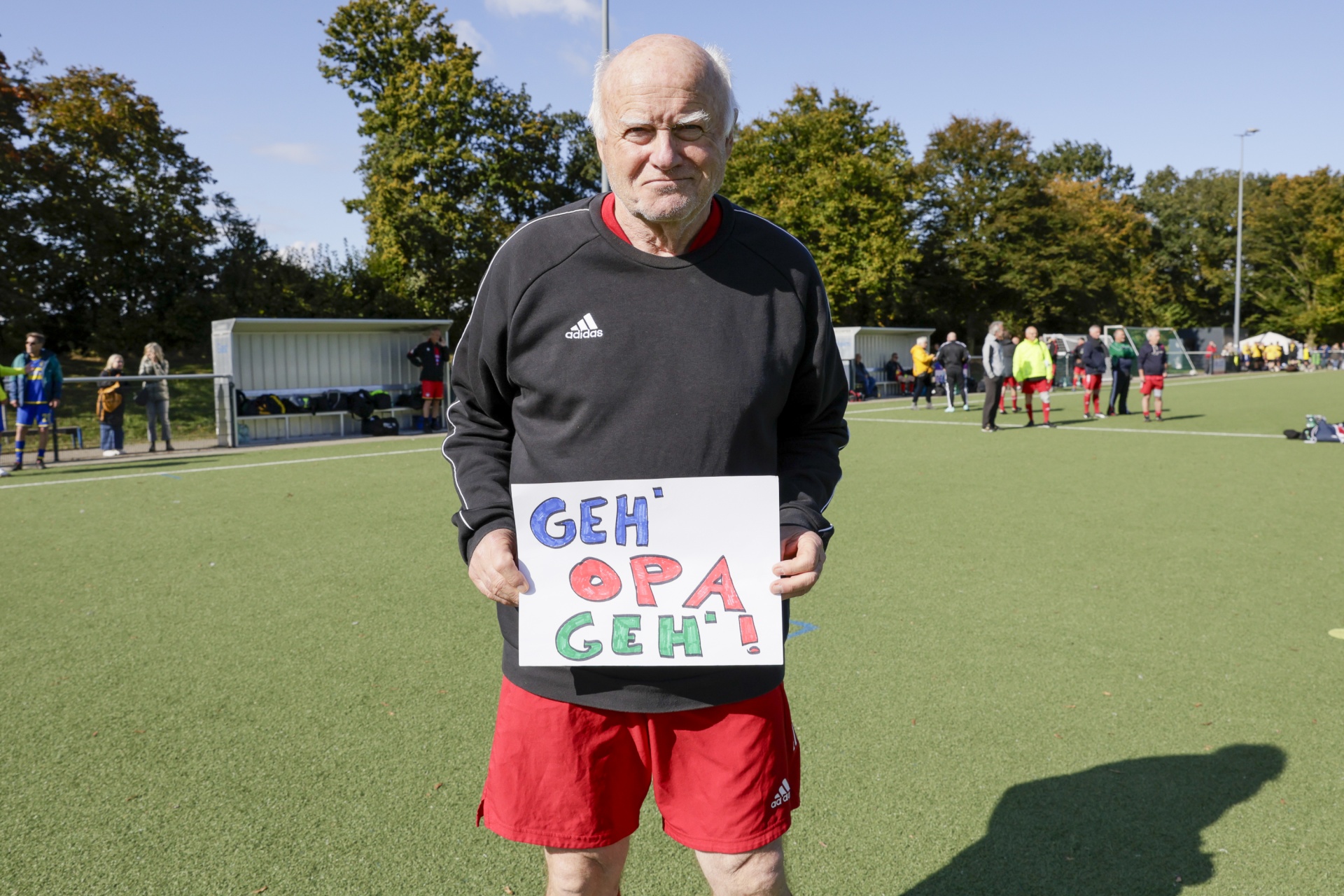 [{"clang_id":1,"value":"Älterer Mann mit grauem Haar und roter Sportbekleidung steht auf einem Fußballfeld. Er hält ein Schild mit der Aufschrift \"GEH' OPA GEH'!\". Im Hintergrund sind weitere Personen und ein Sportplatz zu sehen."}]