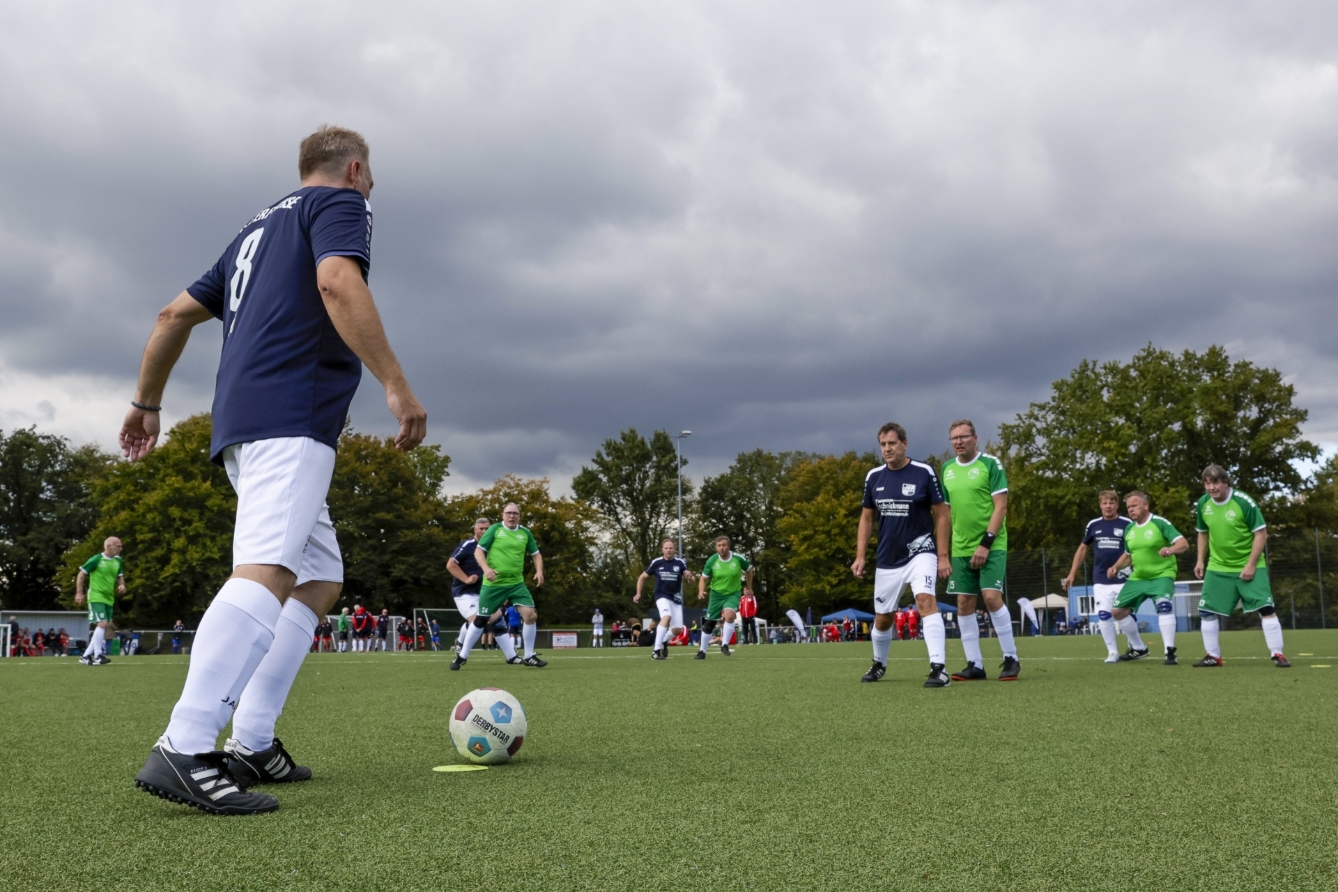 [{"clang_id":1,"value":"Fußballspieler in blauem Trikot schießt auf Ball auf grünem Kunstrasenfeld. Im Hintergrund weitere Spieler in grün und blau sowie Zuschauer. Bewölkter Himmel."}]