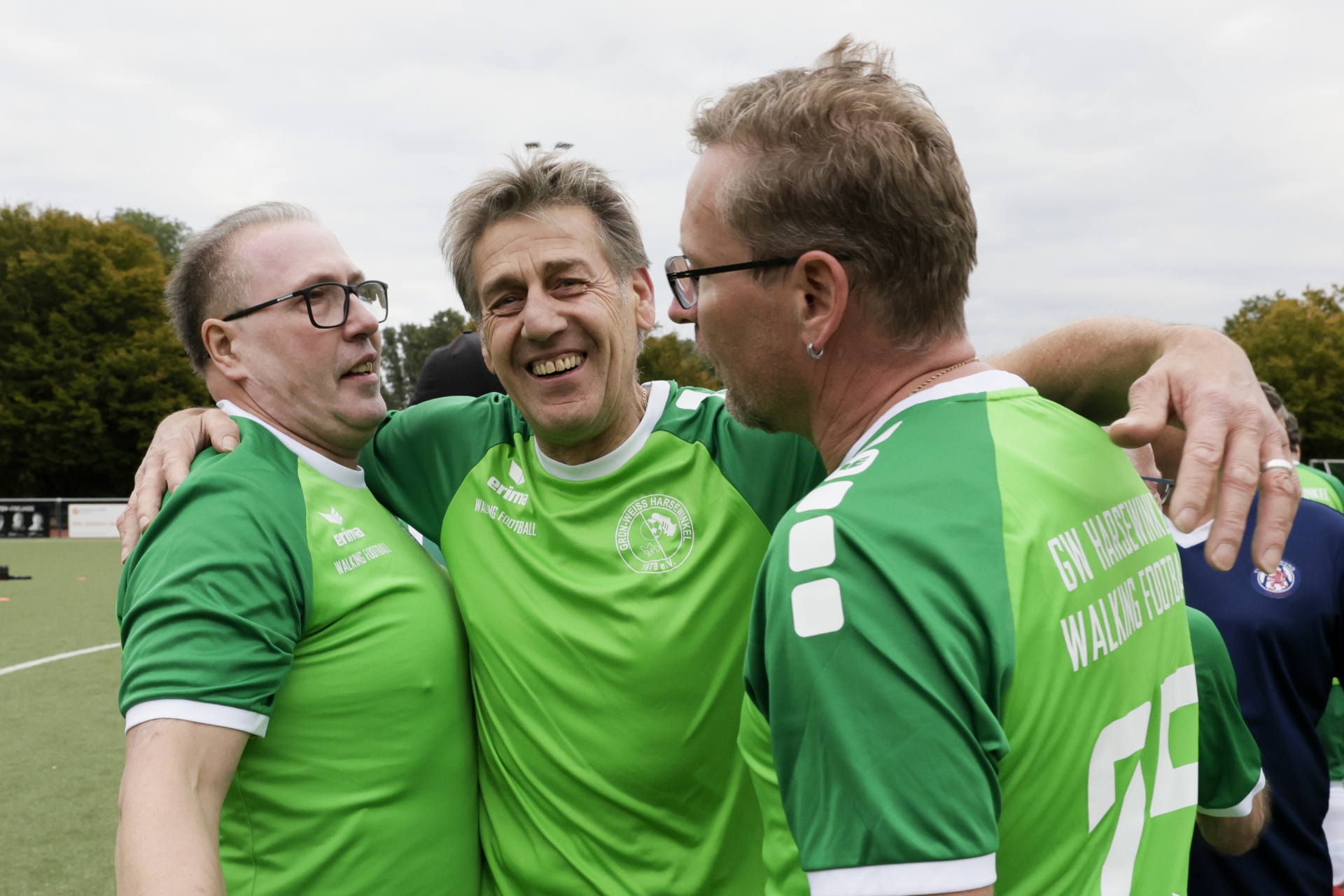 [{"clang_id":1,"value":"Drei Männer in grünen Fußballtrikots umarmen sich lachend auf einem Kunstrasenplatz. Im Hintergrund Bäume und weitere Sportler."}]