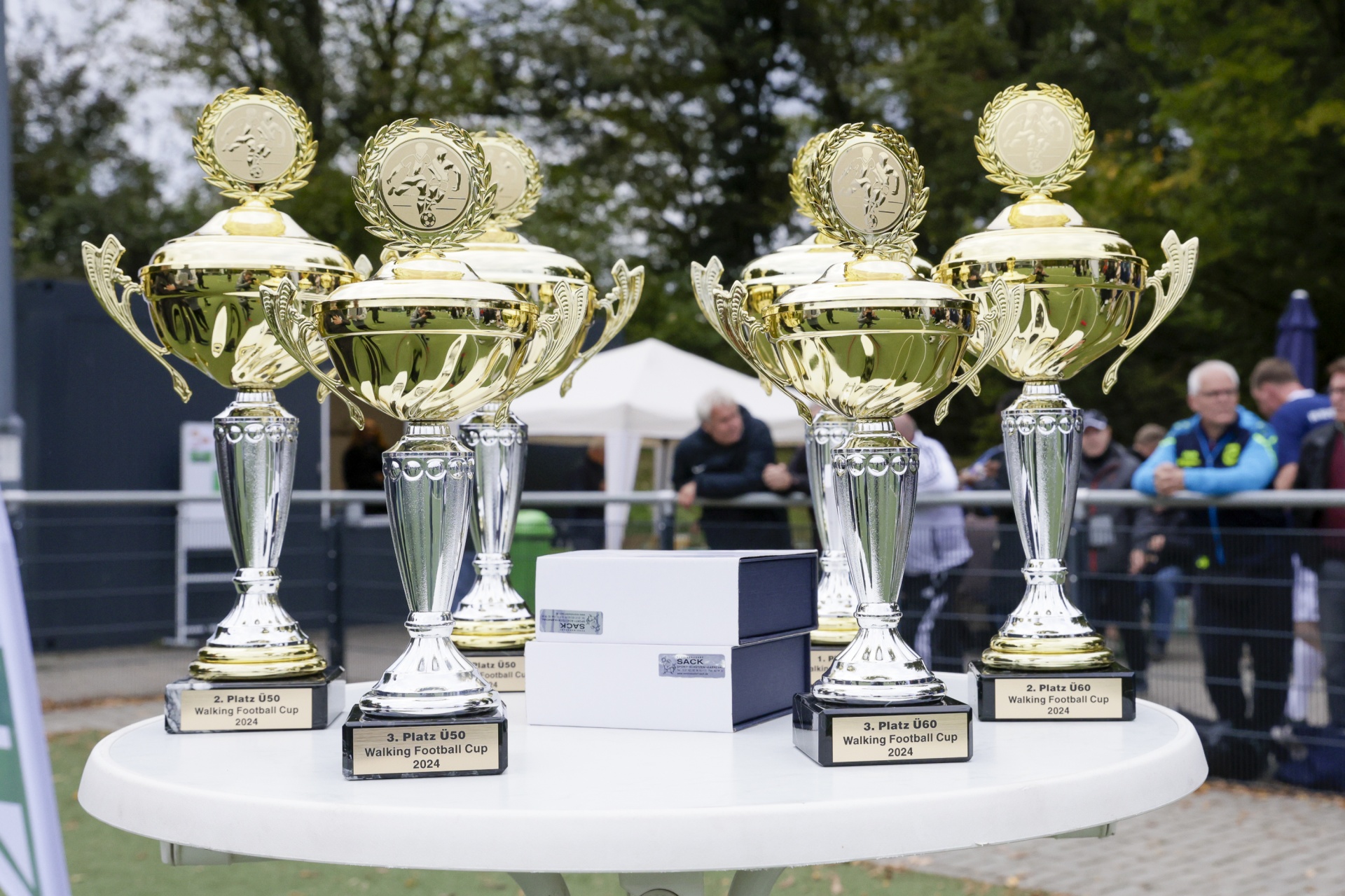 [{"clang_id":1,"value":"Drei goldene Pokale stehen auf einer grauen Platte vor unscharfem Hintergrund mit Zuschauern und grüner Grasfläche. Beschriftungen zeigen \"Platz U50 Walking Football Cup 2024\"."}]
