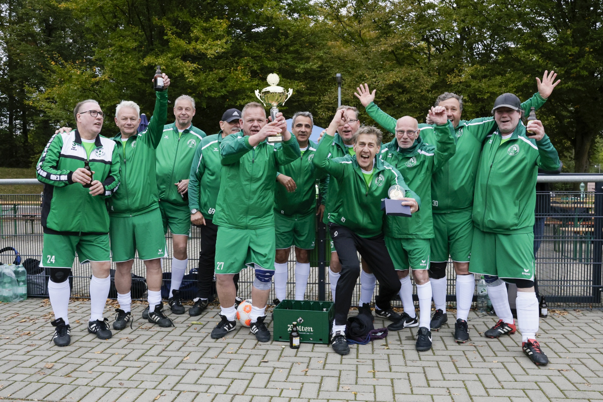 [{"clang_id":1,"value":"Fußballmannschaft in grünen Trikots feiert Sieg mit Pokal. Männer mit hoch erhobenen Armen und Jubelgesten. Im Hintergrund Bäume und eine Absperrung."}]