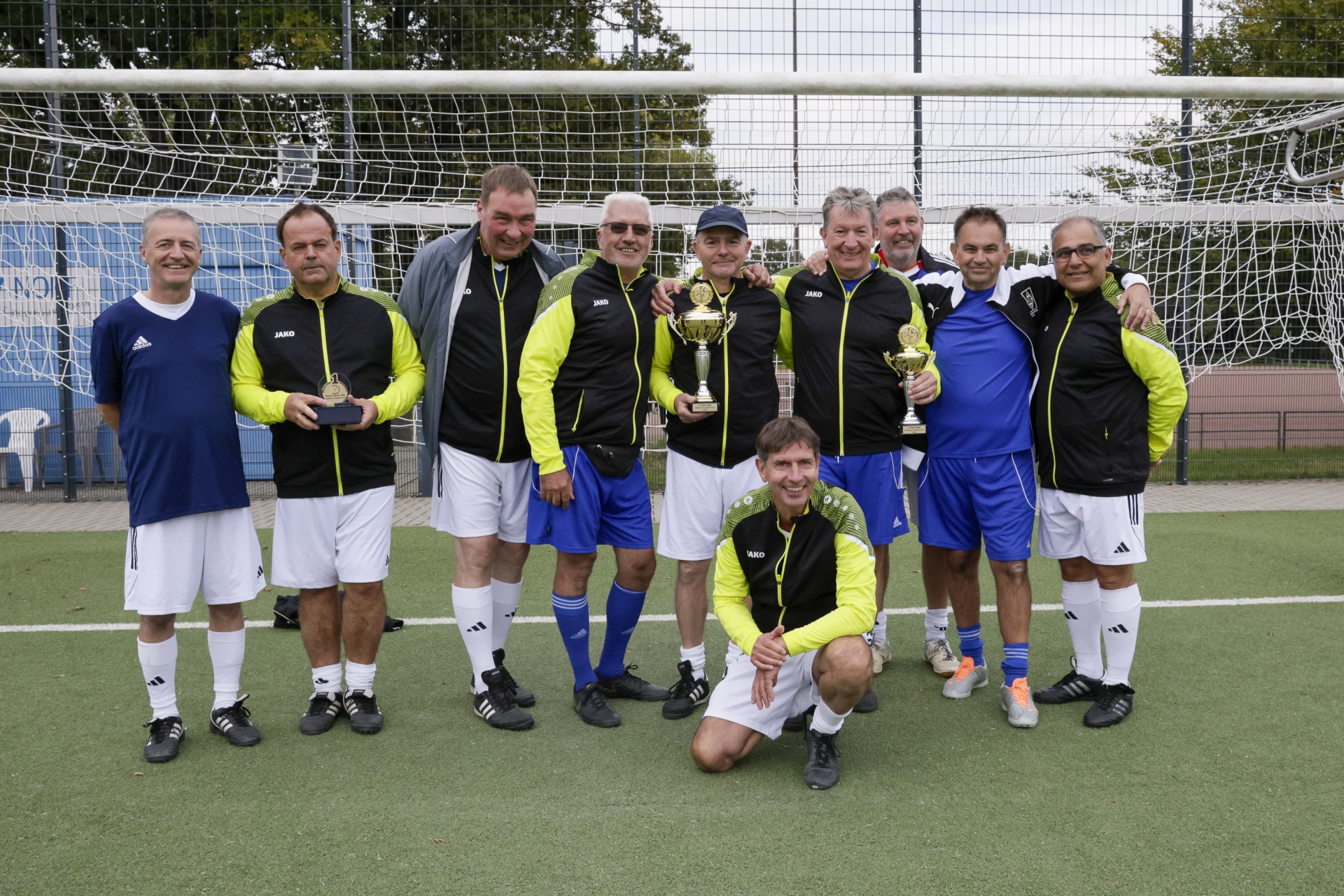 [{"clang_id":1,"value":"Fußballteam posiert mit Pokalen auf grünem Kunstrasen vor einem Fußballtor. Zehn Männer mittleren Alters, sportliche Kleidung."}]
