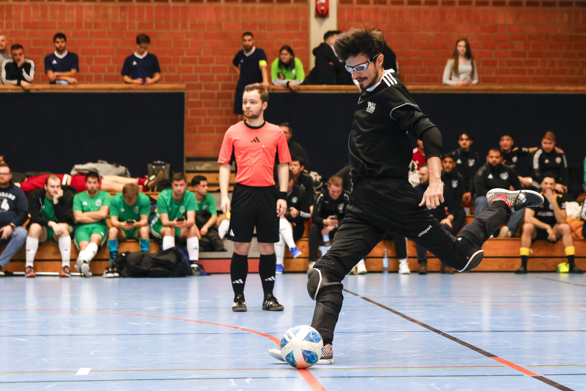 [{"clang_id":1,"value":"Futsal-Spieler mit Brille schießt Ball in Sporthalle. Im Hintergrund Zuschauer und Schiedsrichter."}]