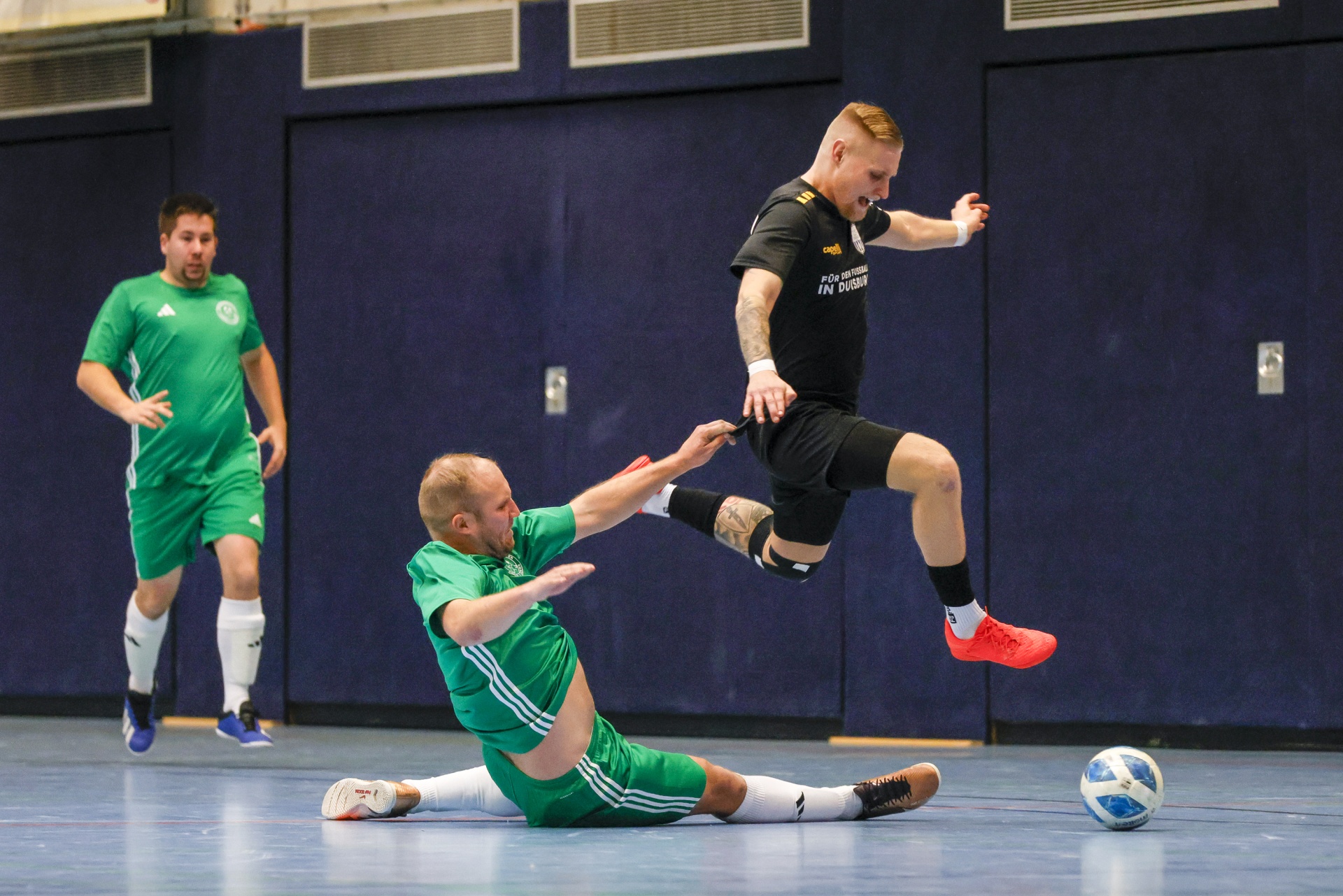 [{"clang_id":1,"value":"Futsalspiel: Zwei Spieler kämpfen um den Ball. Ein Spieler in Grün rutscht, während ein Spieler in Schwarz über ihn springt. Ein dritter Spieler in Grün steht im Hintergrund."}]