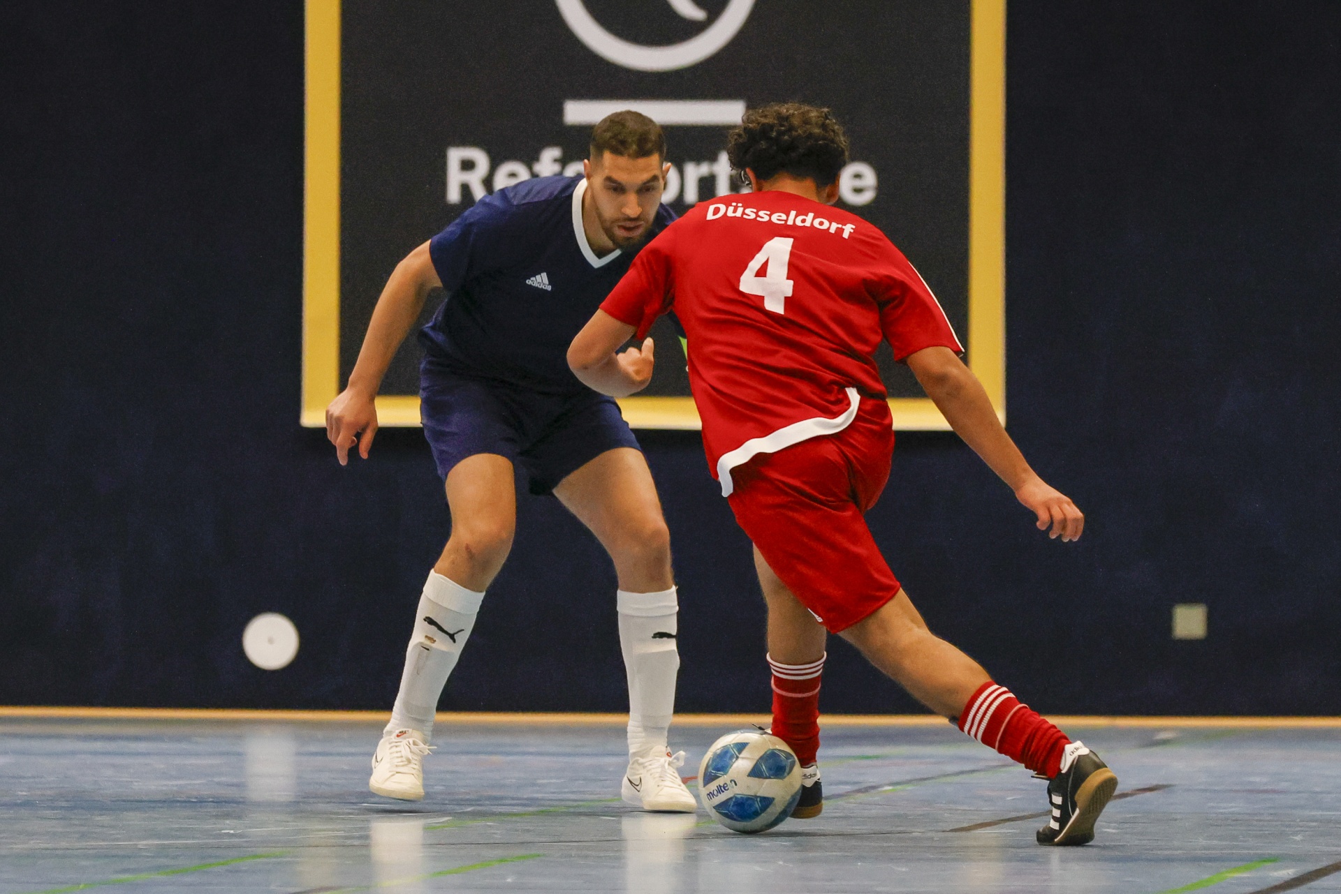 [{"clang_id":1,"value":"Zwei Futsalspieler im Wettkampf. Links ein Spieler in Dunkelblau, rechts in Rot mit Nummer 4, beide fokussiert auf den Ball. Im Hintergrund eine Wand mit Schriftzug."}]