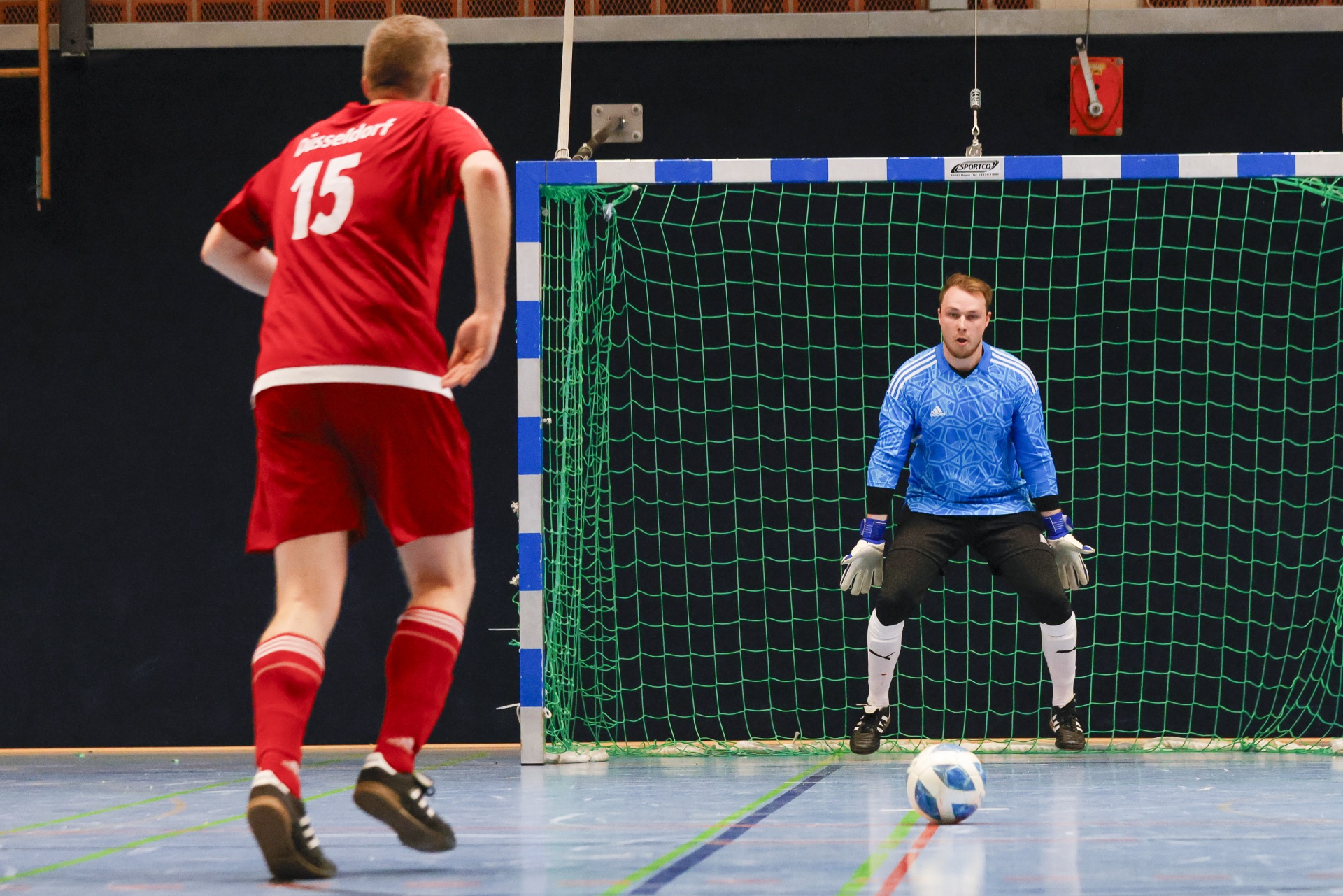 [{"clang_id":1,"value":"Futsal-Spieler mit rotem Trikot (Nr. 15) schießt auf den Torwart in blauem Outfit. Ball zwischen den Spielern. Hallenfußballtor im Hintergrund."}]