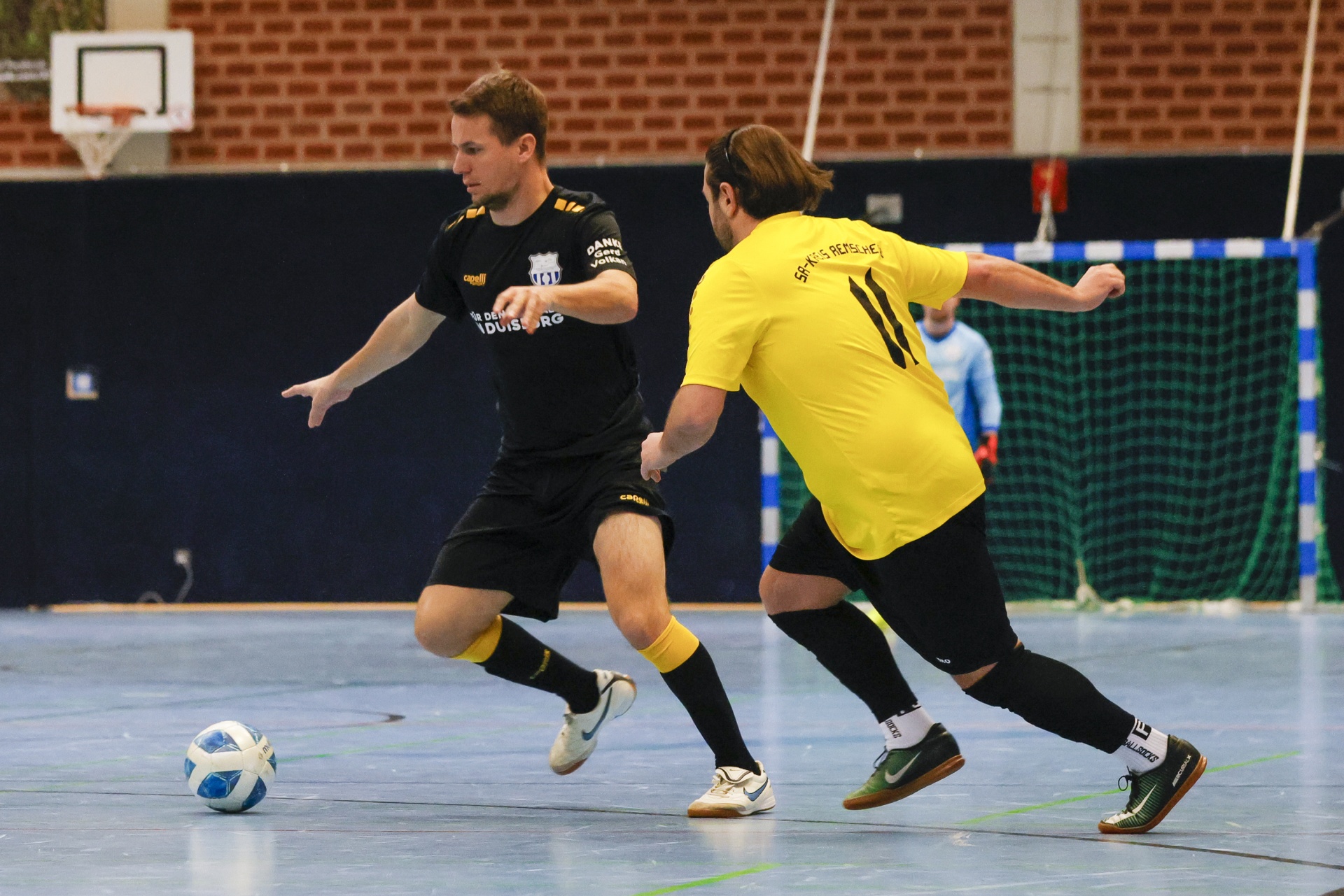[{"clang_id":1,"value":"Zwei Handballspieler in schwarzer und gelber Sportkleidung im Hallensport. Ein Spieler dribbelt den Ball, der andere verfolgt ihn. Im Hintergrund ein Tor und eine Tribüne."}]