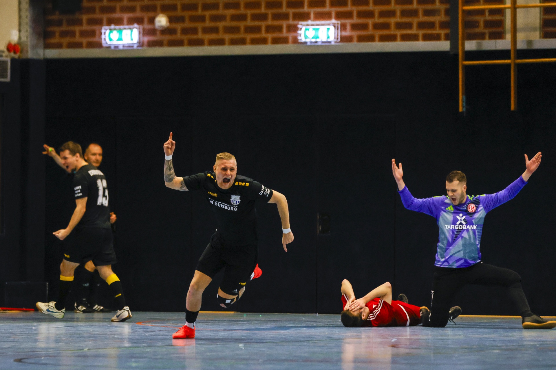 [{"clang_id":1,"value":"Handballspiel: Spieler in schwarzen und violetten Trikots feiern, ein Spieler liegt am Boden. Im Hintergrund eine Sporthalle."}]