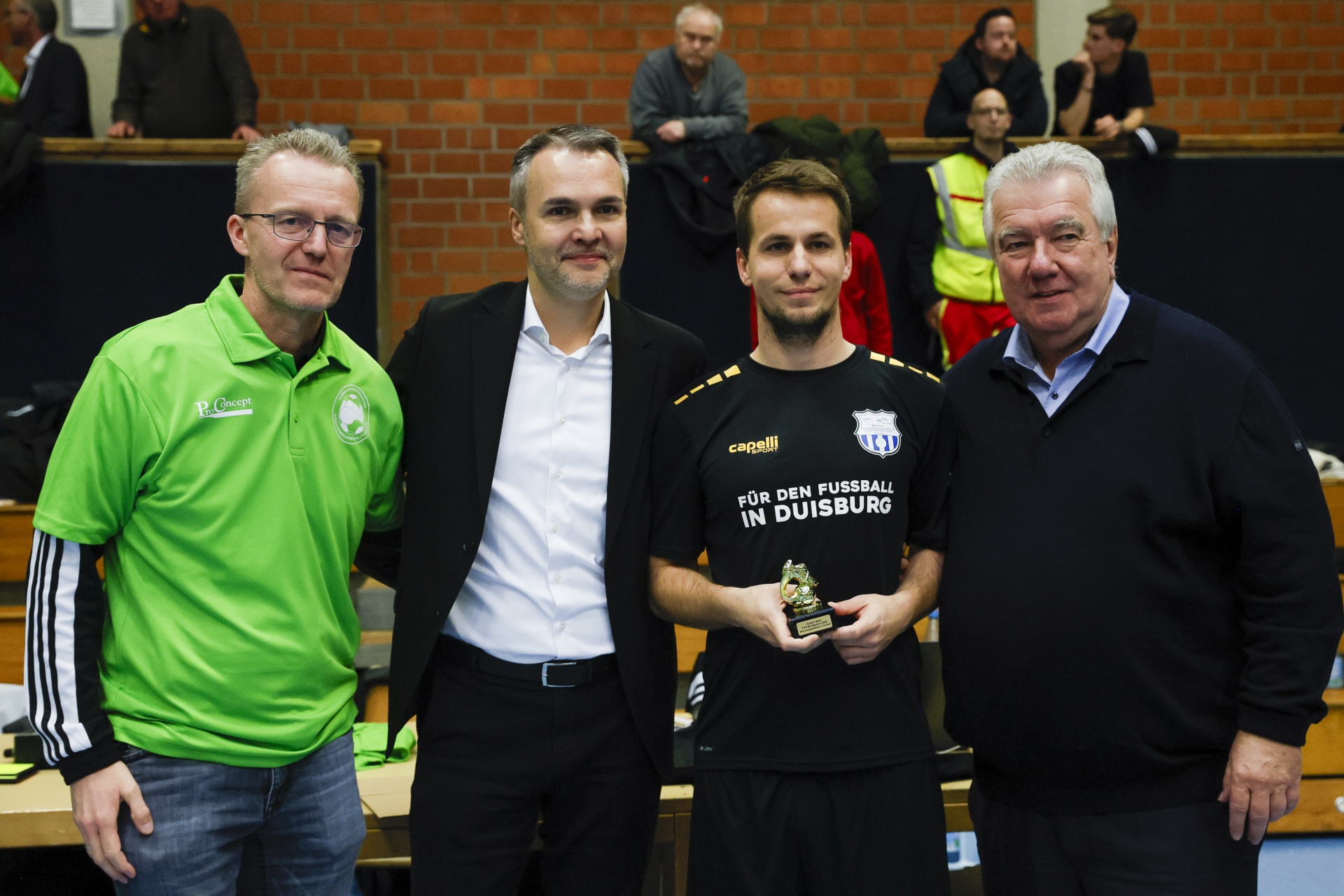 [{"clang_id":1,"value":"Vier Männer stehen nebeneinander in einer Sporthalle. Links trägt einer grünes Poloshirt, in der Mitte zwei Männer im Anzug, rechts einer grauen Pullover. Ein Mann hält eine kleine Trophäe."}]