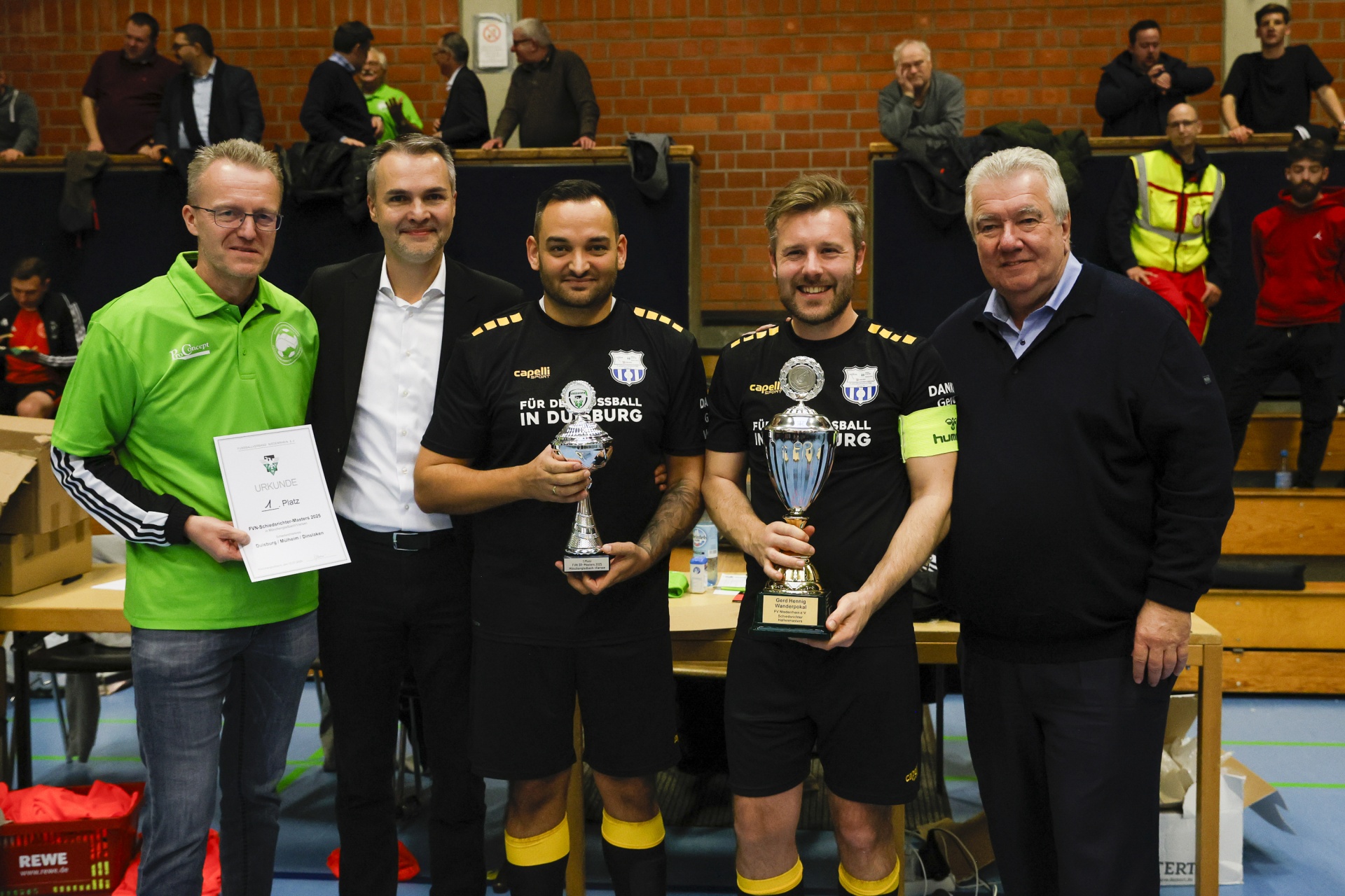 [{"clang_id":1,"value":"Fünf Männer stehen nebeneinander in einer Sporthalle. Zwei halten Trophäen und Urkunden. Kleidung: Sportshirts, Pullover, Jacke. Hintergrund: Zuschauer und eine Wand mit Holzvertäfelung."}]