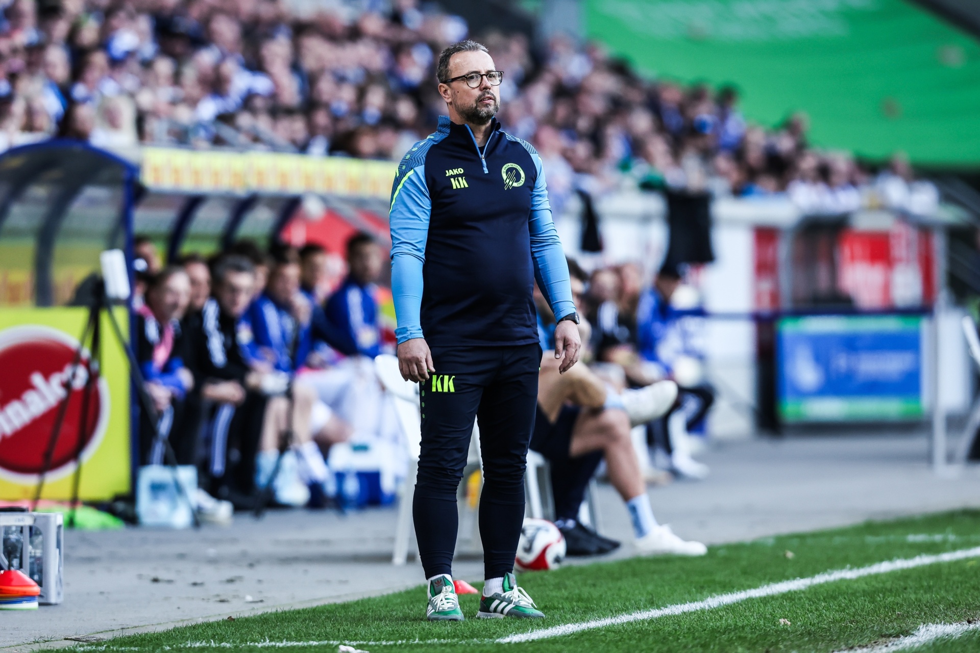 [{"clang_id":1,"value":"Mann mit Bart und Brille, Trainingsanzug, steht am Spielfeldrand vor vollbesetzten Tribünen eines Fußballstadions. Grüner Rasen im Vordergrund."}]