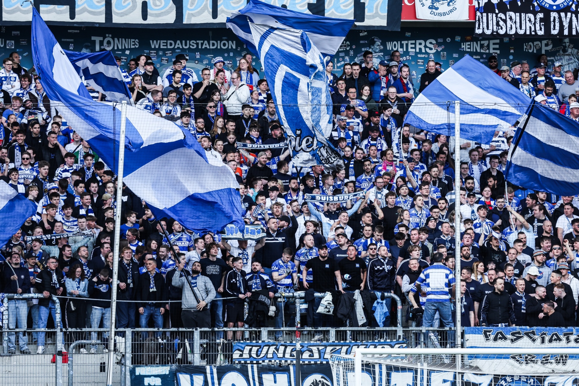 [{"clang_id":1,"value":"Fußballstadion: Dichte Menschenmenge mit blau-weißen Fahnen und Bannern. Im Hintergrund Werbebanner."}]