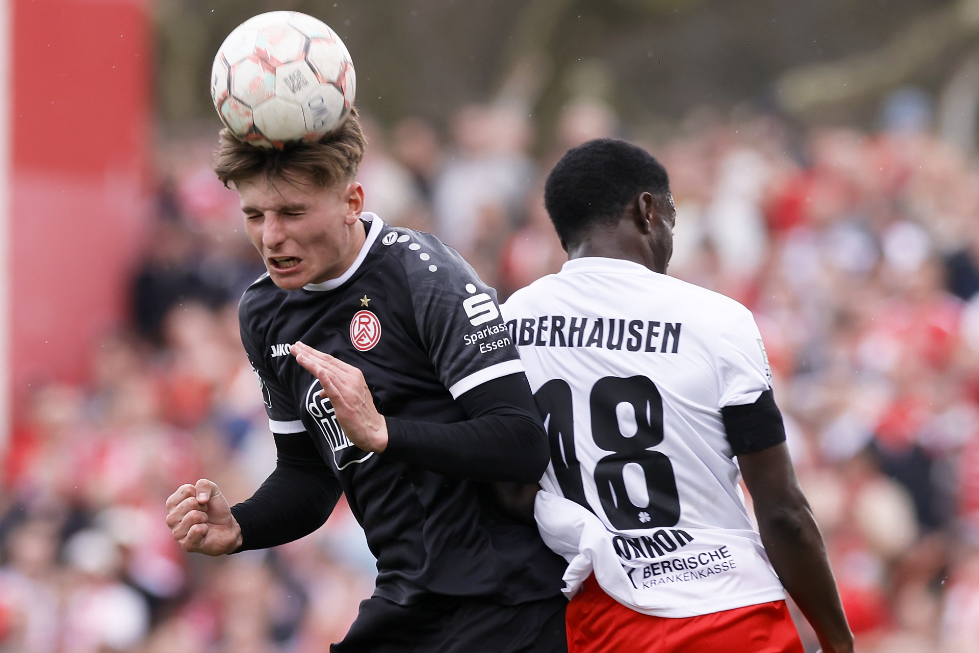 [{"clang_id":1,"value":"Zwei Fußballspieler im Spiel. Ein Spieler mit rotem Trikot köpft den Ball, während ein anderer Spieler mit schwarzem Trikot und Nummer 18 dahinter steht. Stadion im Hintergrund."}]