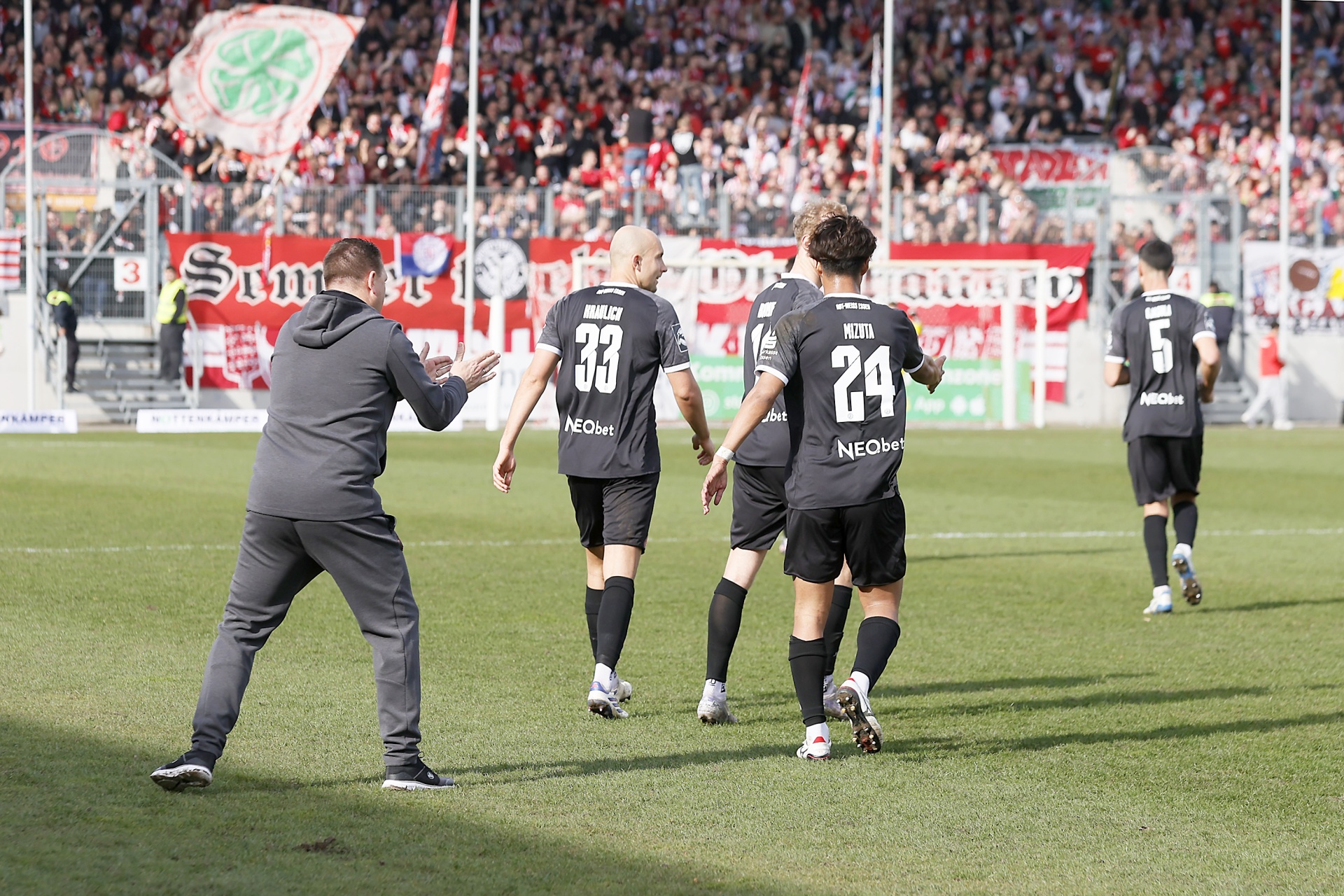 [{"clang_id":1,"value":"Fußballspiel: Vier Spieler in schwarzen Trikots laufen auf dem Rasen. Ein Trainer mit Jacke und Hose steht links, die Arme gehoben. Im Hintergrund Zuschauer."}]