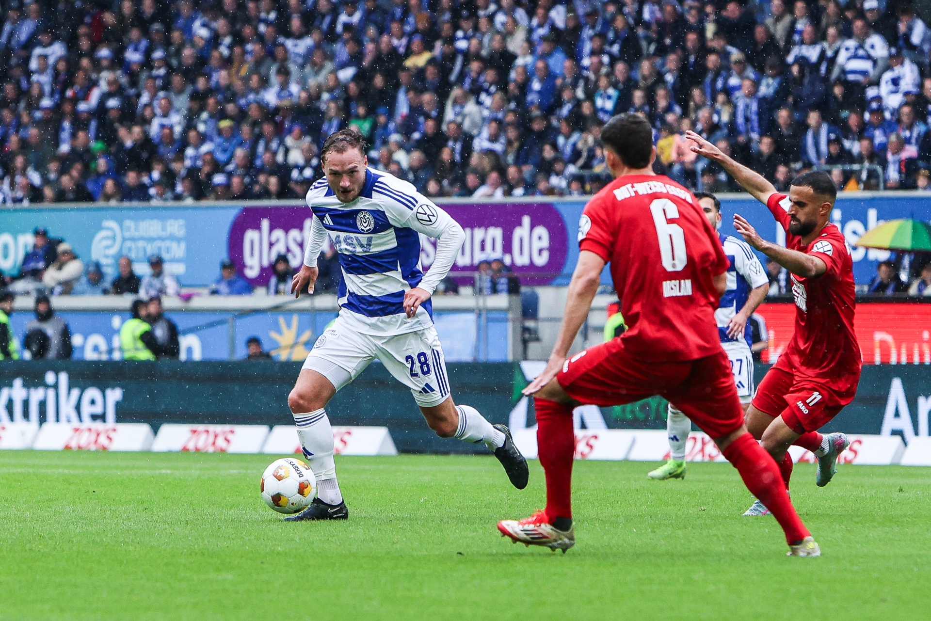 [{"clang_id":1,"value":"Fußballspiel: Spieler mit Nummer 28 in blauem Trikot schießt auf das Tor, während zwei Gegenspieler in roten Trikots ihm entgegenkommen. Stadion im Hintergrund."}]