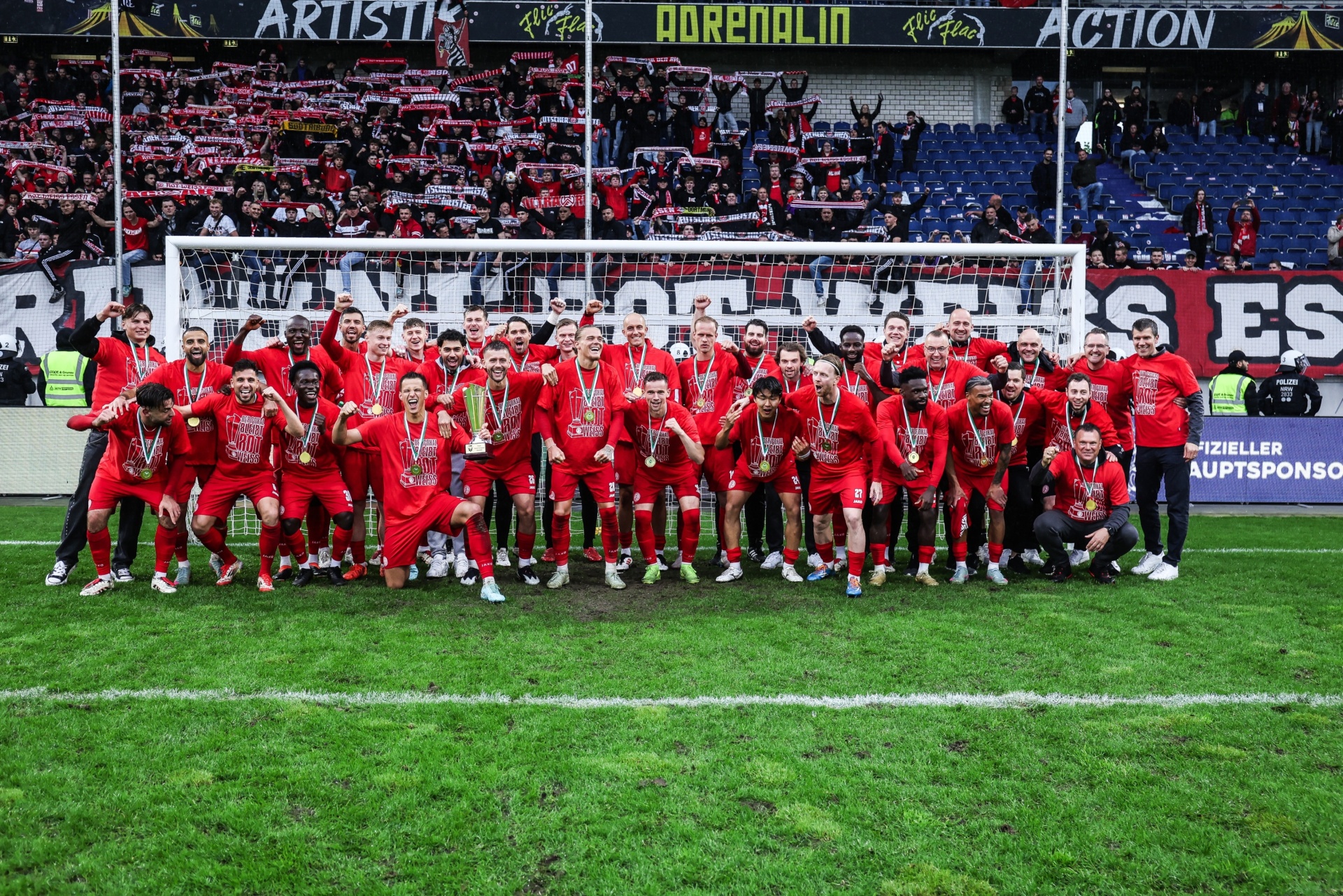 [{"clang_id":1,"value":"Fußballmannschaft in roten Trikots kniet vor dem Tor und feiert. Im Hintergrund: gefülltes Stadion mit Zuschauern. Banner mit der Aufschrift \"ARTISTIC\", \"ADRENALIN\", \"ACTION\" sind zu sehen."}]