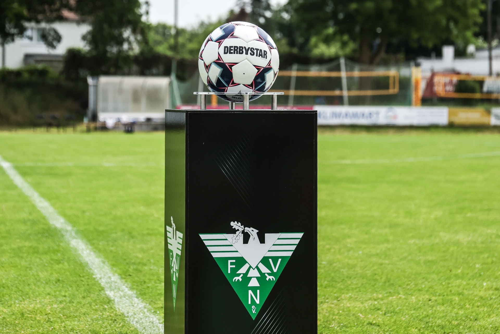 [{"clang_id":1,"value":"Fußball auf Säule: Ein Derby Star Fußball ruht auf einer schwarzen Säule mit grün-weißem Wappen (F.V.N.N.) vor einem Fußballfeld."}]