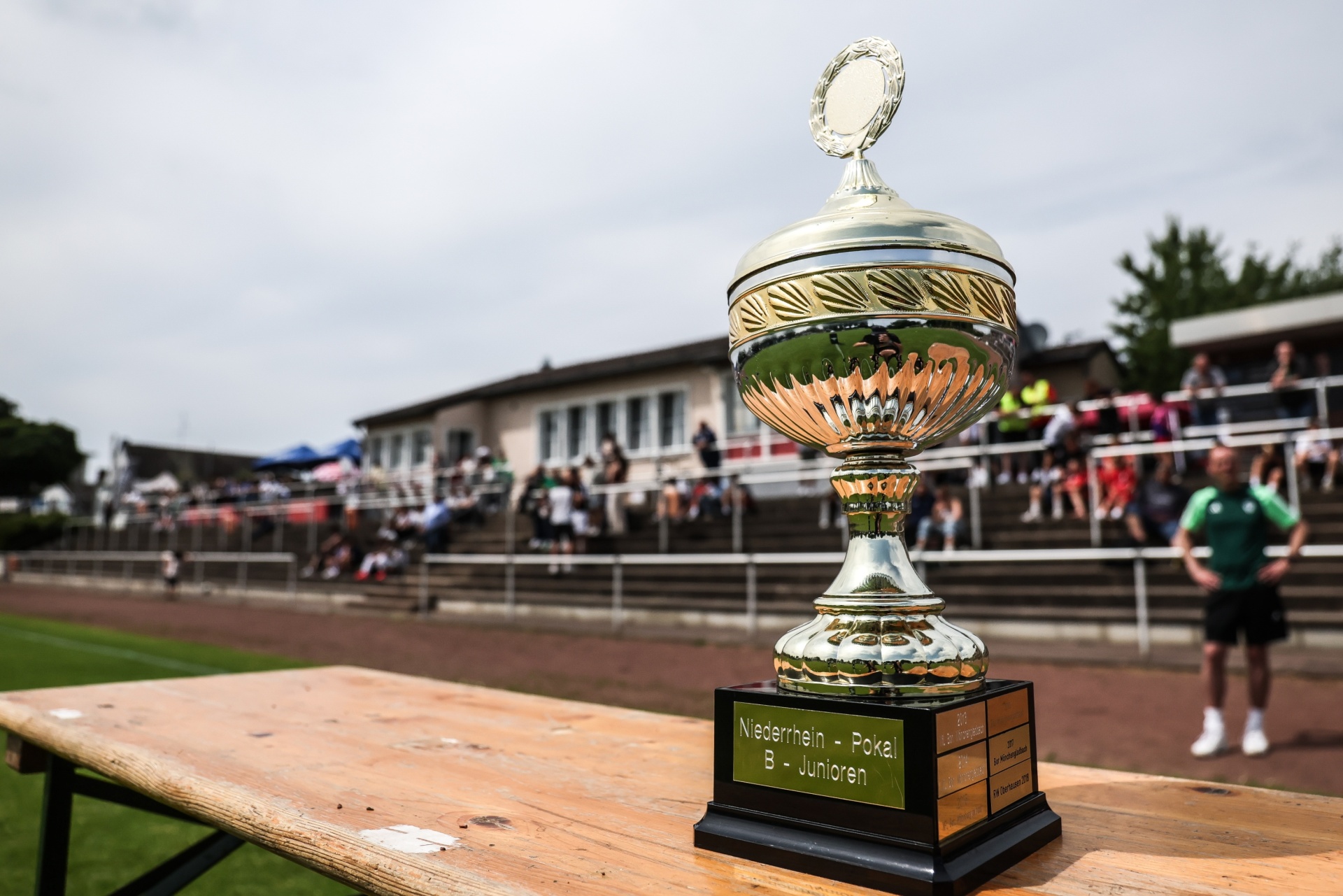 [{"clang_id":1,"value":"Pokal auf einer Holzbank vor einem Fußballstadion mit Zuschauern. Beschriftung am Pokal: \"Niederrhein - Pokal B - Junioren\"."}]