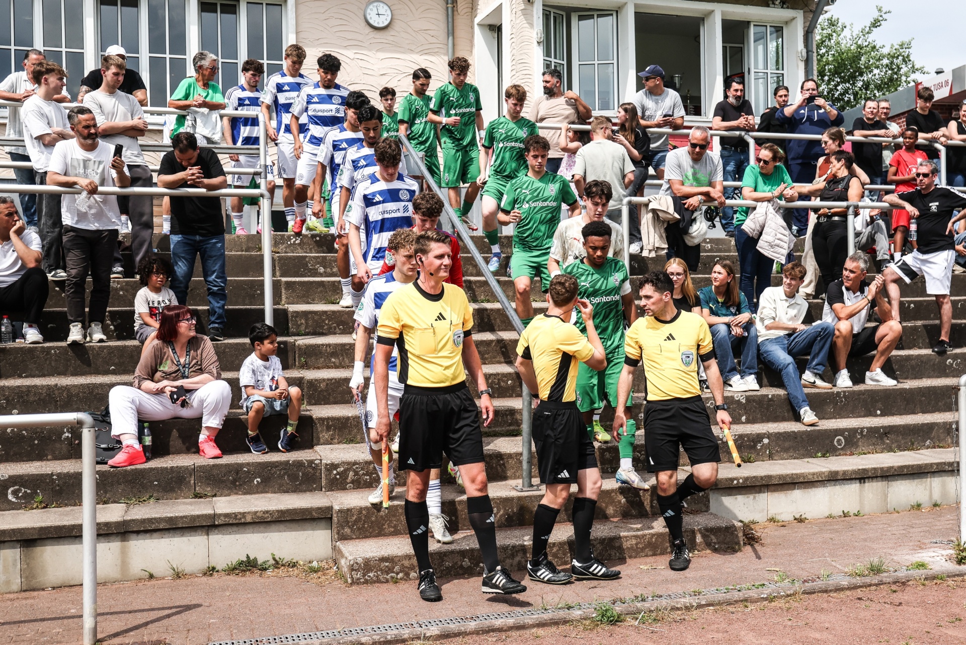[{"clang_id":1,"value":"Fußballspiel: Schiedsrichter in Gelb-Schwarz betreten den Platz. Im Hintergrund Zuschauerreihen mit Fans in Vereinsfarben Grün und Blau-Weiß."}]