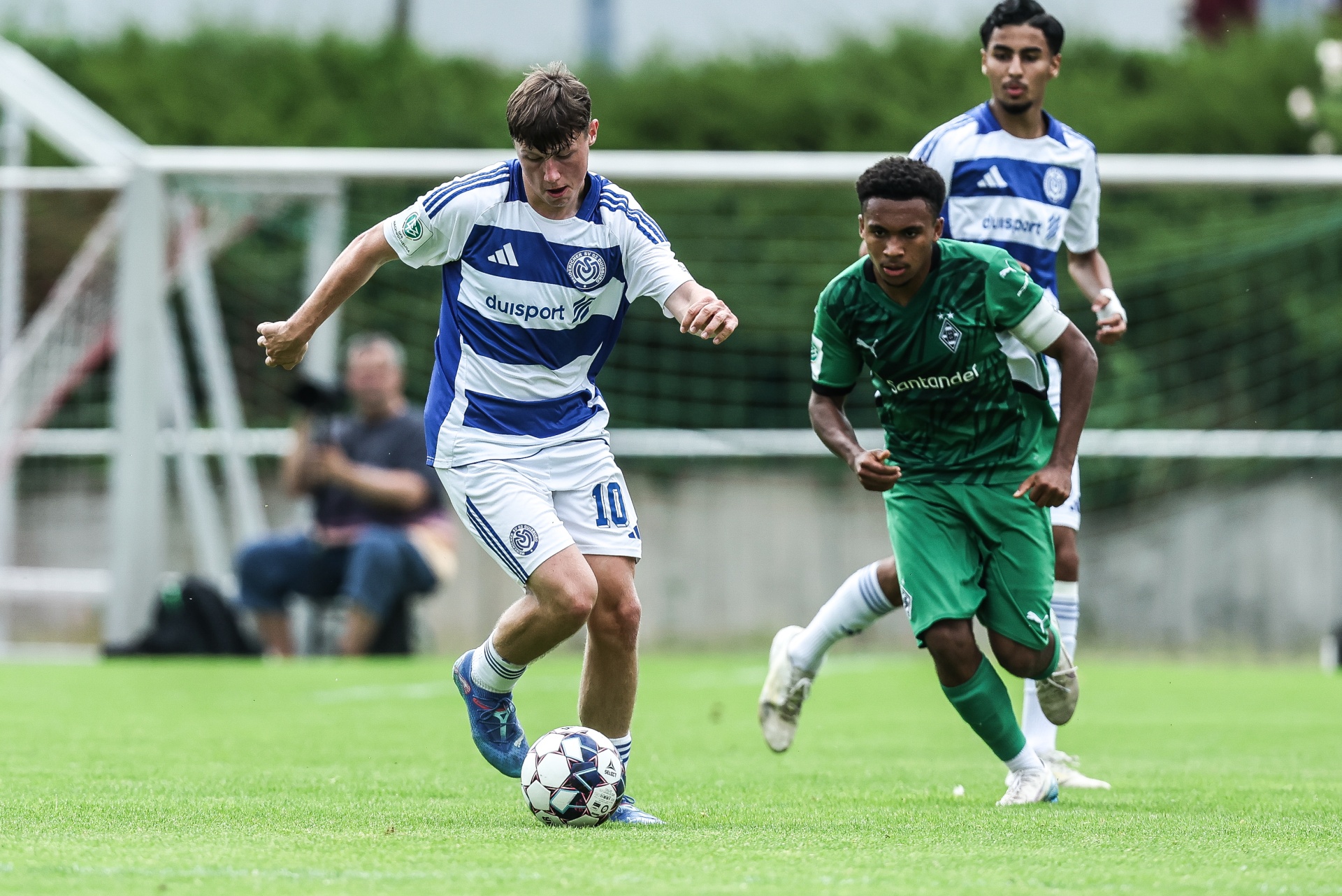 [{"clang_id":1,"value":"Zwei Fußballspieler auf grünem Rasen. Links Spieler mit blau-weißem Trikot und Ball, rechts Spieler mit grünem Trikot im Wettlauf. Fußballtor im Hintergrund."}]