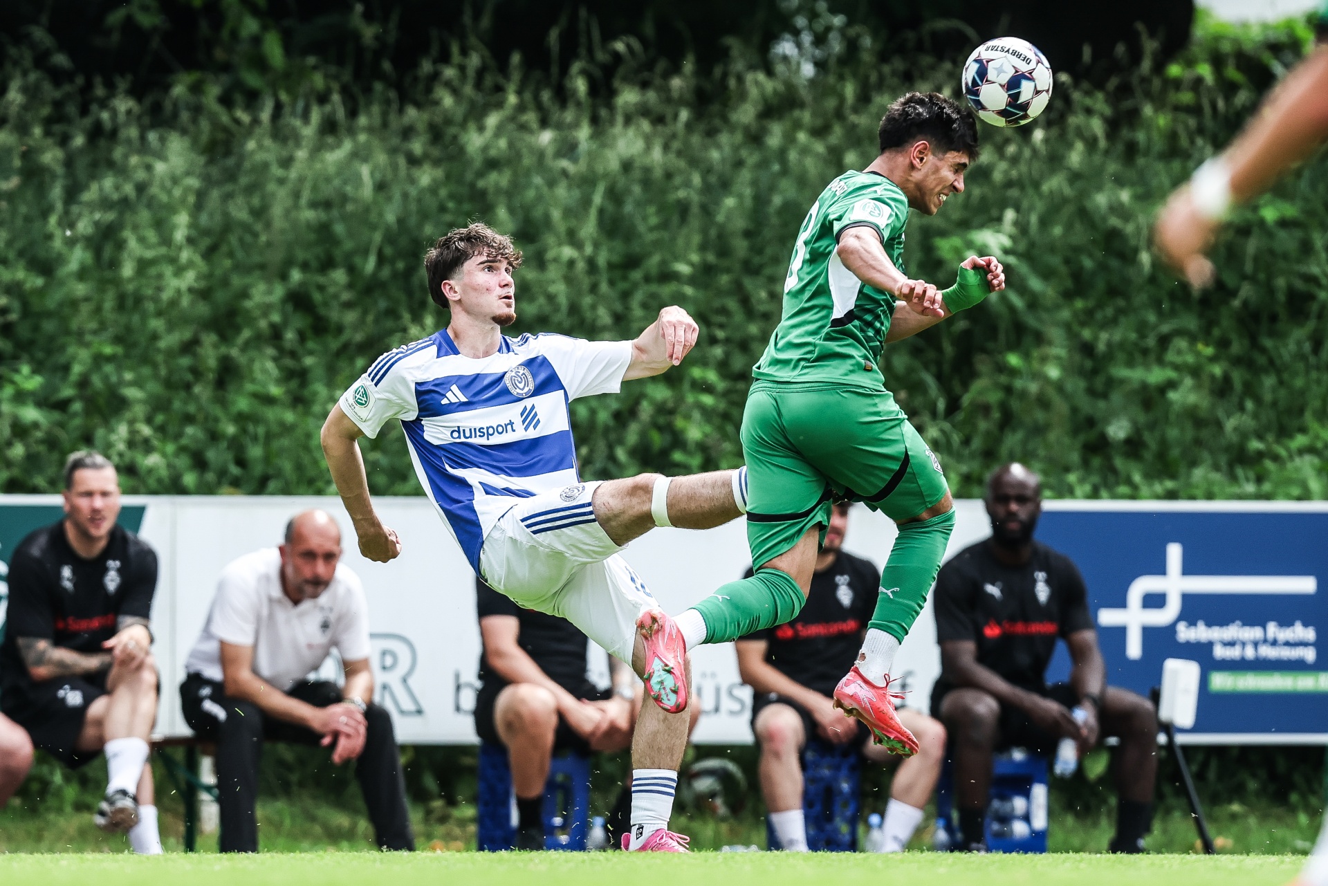 [{"clang_id":1,"value":"Zwei Fußballspieler im Zweikampf: Ein Spieler in blau-weißem Trikot versucht, den Ball gegen einen Spieler in grünem Trikot zu treten, der in der Luft ist. Im Hintergrund Zuschauer und Bäume."}]
