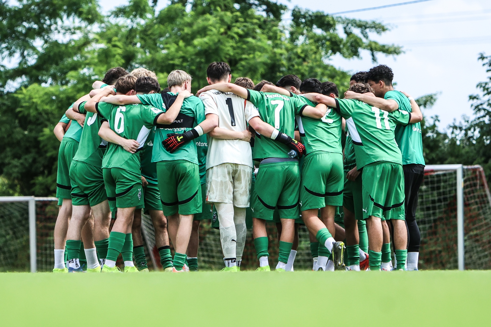 [{"clang_id":1,"value":"Fußballmannschaft in grünen Trikots steht im Kreis und umarmt sich auf einem Fußballfeld. Im Hintergrund ein Fußballtor und Bäume."}]