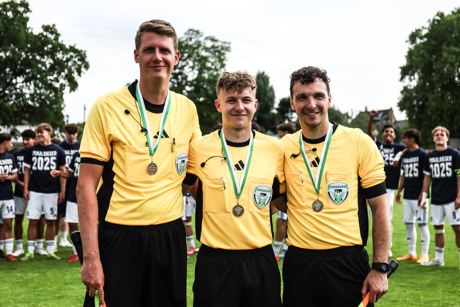 [{"clang_id":1,"value":"Drei Schiedsrichter in gelben Trikots und schwarzen Shorts mit Medaillen auf dem Fußballplatz. Im Hintergrund stehen Spieler in blauen Trikots."}]