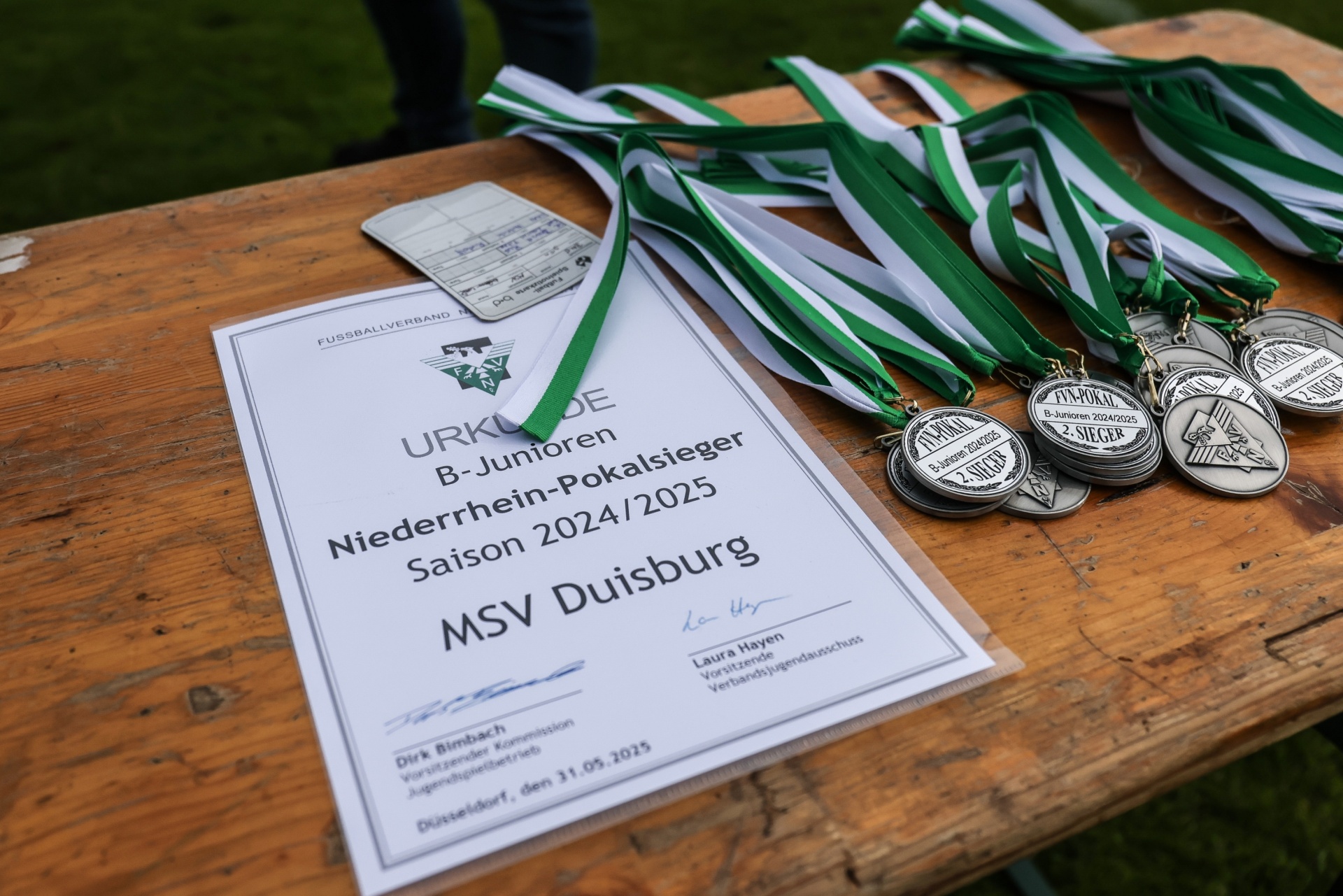 [{"clang_id":1,"value":"Fußball-Pokal-Urkunde für MSV Duisburg B-Junioren, Saison 2024/2025. Medaillen und grüne Bänder liegen darauf. Holzbank im Hintergrund."}]