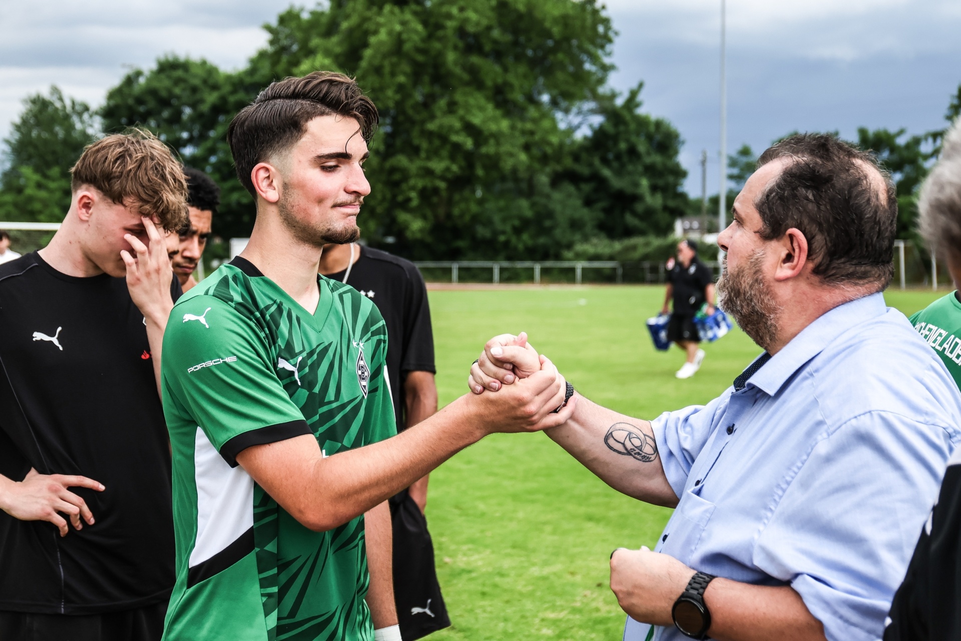 [{"clang_id":1,"value":"Zwei Männer begrüßen sich feierlich mit einer Faust. Im Hintergrund Fußballspieler auf einem grünen Rasenfeld unter grauem Himmel."}]