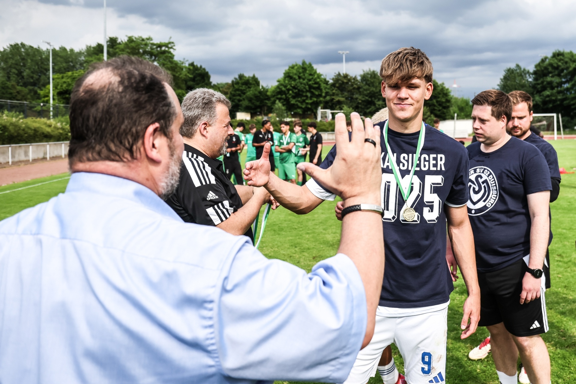 [{"clang_id":1,"value":"Fußballspieler mit Trikotnummer 9 erhält High-Five von Trainer und Begleitern auf Fußballfeld. Im Hintergrund weitere Spieler und Zuschauer."}]