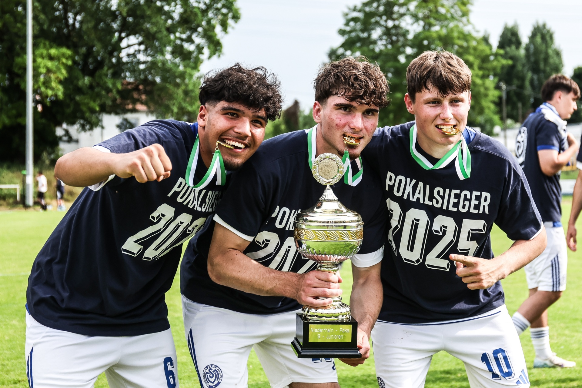 [{"clang_id":1,"value":"Drei junge Fußballspieler mit Medaillen um den Hals umarmen einen goldenen Pokal. Sie tragen dunkelblaue Trikots mit der Aufschrift \"POKALSieger 2025\". Im Hintergrund weitere Spieler und Grünflächen."}]