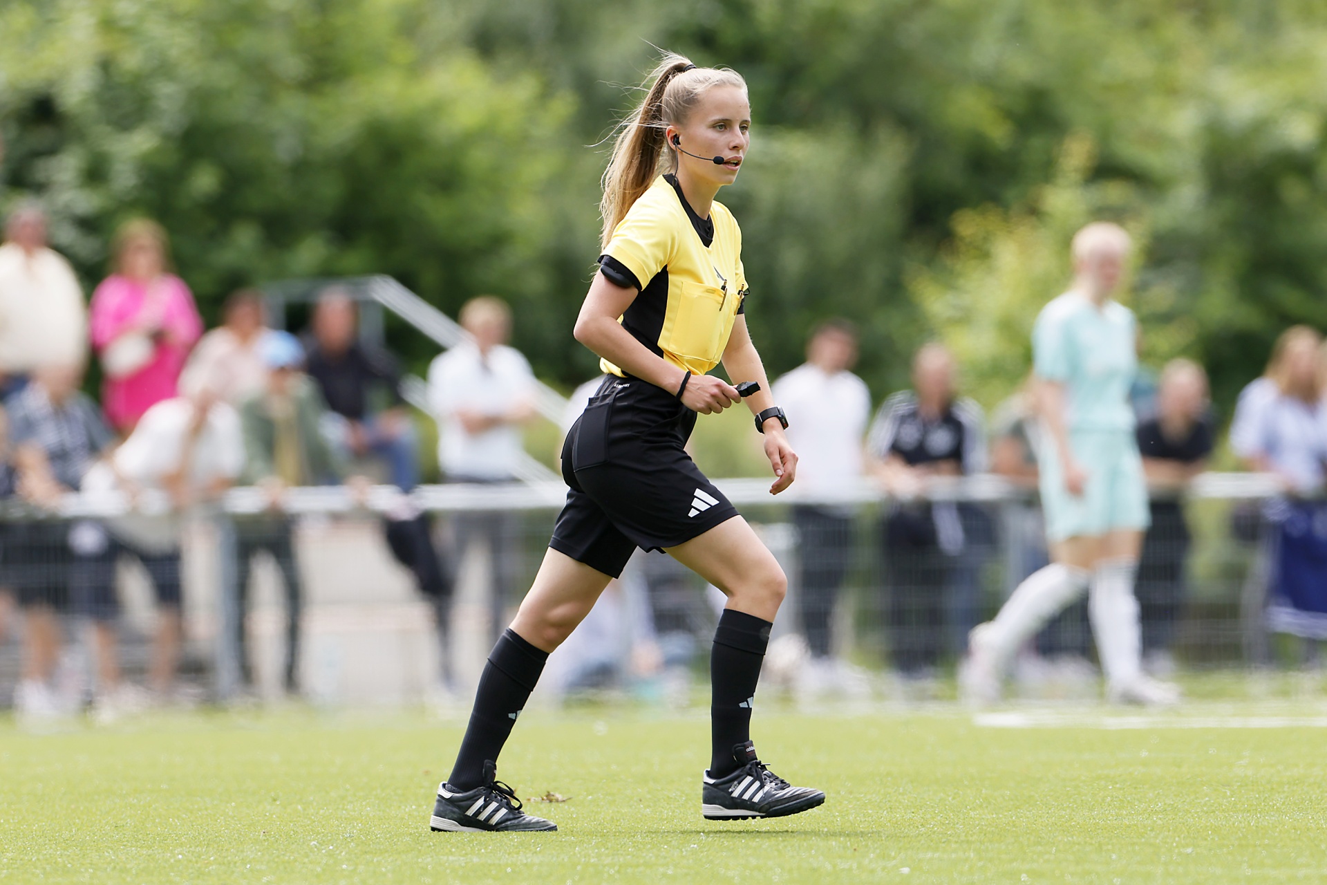 [{"clang_id":1,"value":"Fußballschiedsrichterin in schwarzer Hose und gelbem Trikot auf grünem Rasen. Sie trägt Headset und Armbanduhr. Im Hintergrund Zuschauer und weitere Spieler."}]
