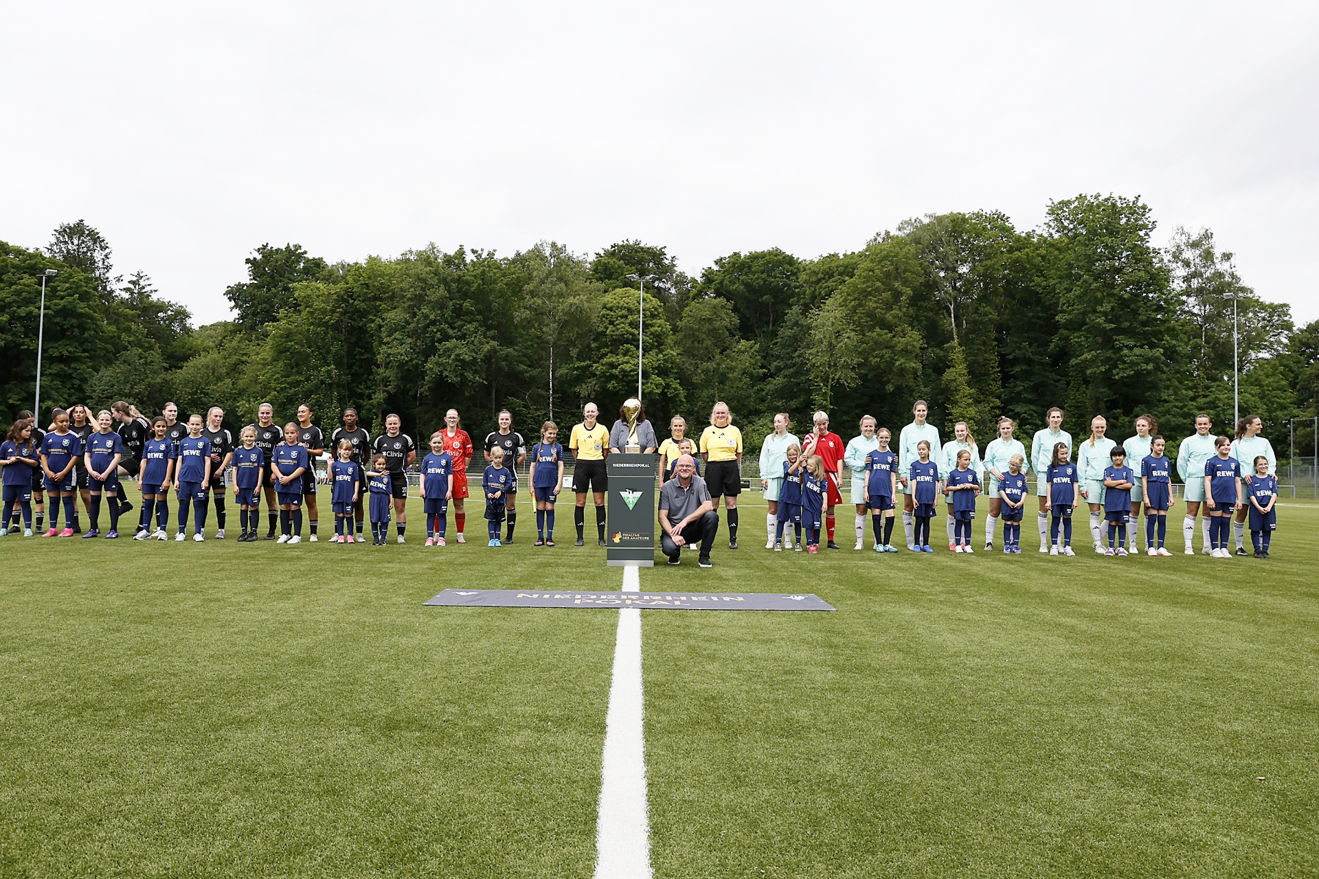 [{"clang_id":1,"value":"Fußballmannschaften und Funktionäre stehen auf einem grünen Rasenfeld vor einem dunklen Denkmal und Baumreihe. Eine weiße Linie teilt den Platz."}]
