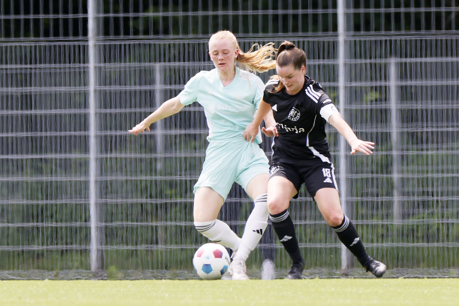 [{"clang_id":1,"value":"Zwei Fußballspielerinnen im Spiel. Eine trägt hellblaues Trikot, die andere schwarz-weißes. Sie kämpfen um den Ball auf einem grünen Rasenplatz vor einem Metallzaun."}]