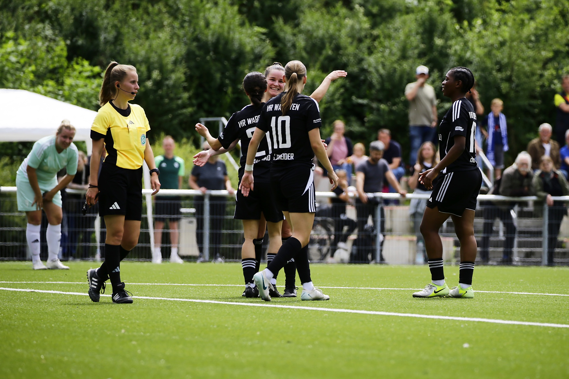 [{"clang_id":1,"value":"Fußballspiel: Spielerinnen in schwarz-weißen Trikots jubeln mit der Schiedsrichterin. Im Hintergrund Zuschauer und grüne Bäume."}]