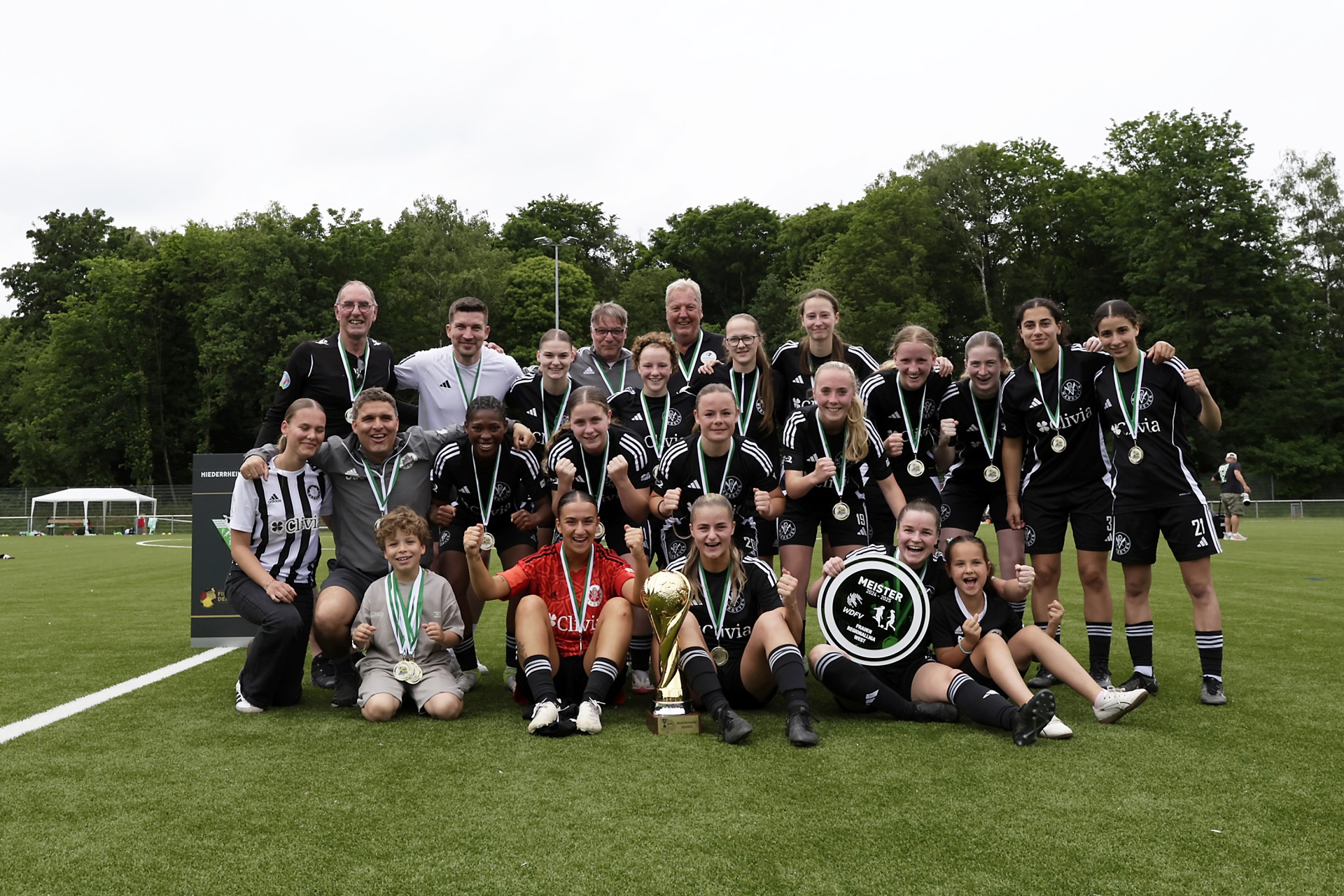[{"clang_id":1,"value":"Fußballmannschaft mit Trainer und Betreuern posiert auf Rasenfeld mit Pokal und Medaillen. Spieler tragen schwarz-weiße Trikots. Bäume im Hintergrund."}]