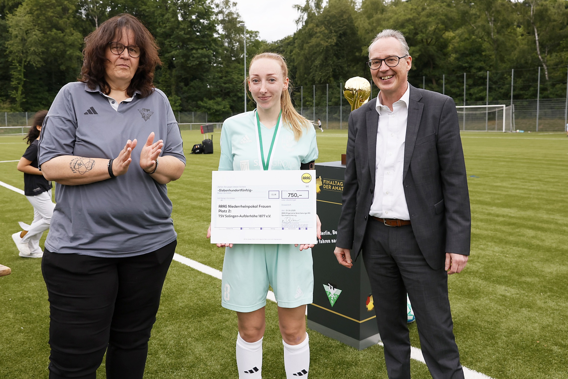 [{"clang_id":1,"value":"Drei Personen überreichen einen symbolischen Scheck auf einem Fußballfeld. Links eine Frau, in der Mitte eine junge Sportlerin und rechts ein Mann im Anzug. Im Hintergrund ein Pokal und ein Zaun."}]