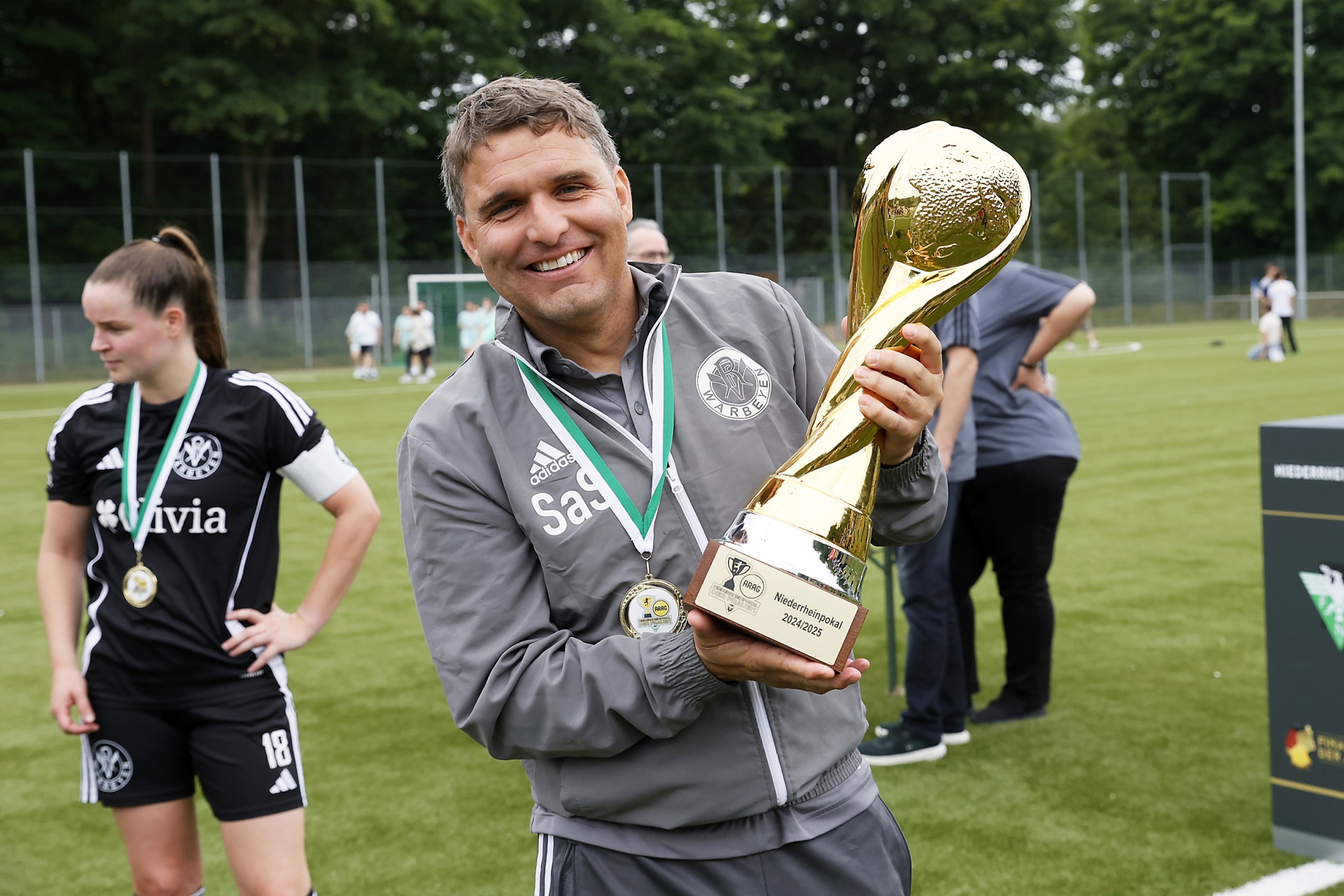 [{"clang_id":1,"value":"Mann mit Jacke hält Pokal im Fußballstadion. Links eine Frau mit Sportkleidung und Medaille. Im Hintergrund grüne Bäume und Spielfeld."}]