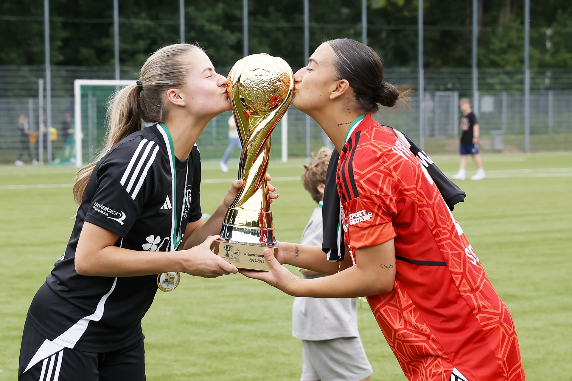 [{"clang_id":1,"value":"Zwei Fußballspielerinnen küssen sich vor dem Hintergrund eines Fußballfeldes. Die eine trägt ein schwarzes, die andere ein rotes Trikot. Beide halten einen goldenen Pokal."}]