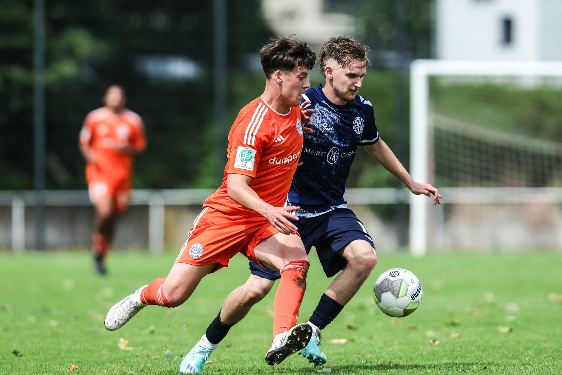 [{"clang_id":1,"value":"Zwei Fußballspieler im Zweikampf auf grünem Rasen. Links trägt ein Spieler orange-blaues Trikot, rechts dunkelblaues. Im Hintergrund weitere Spieler und eine Zuschauertribüne."}]