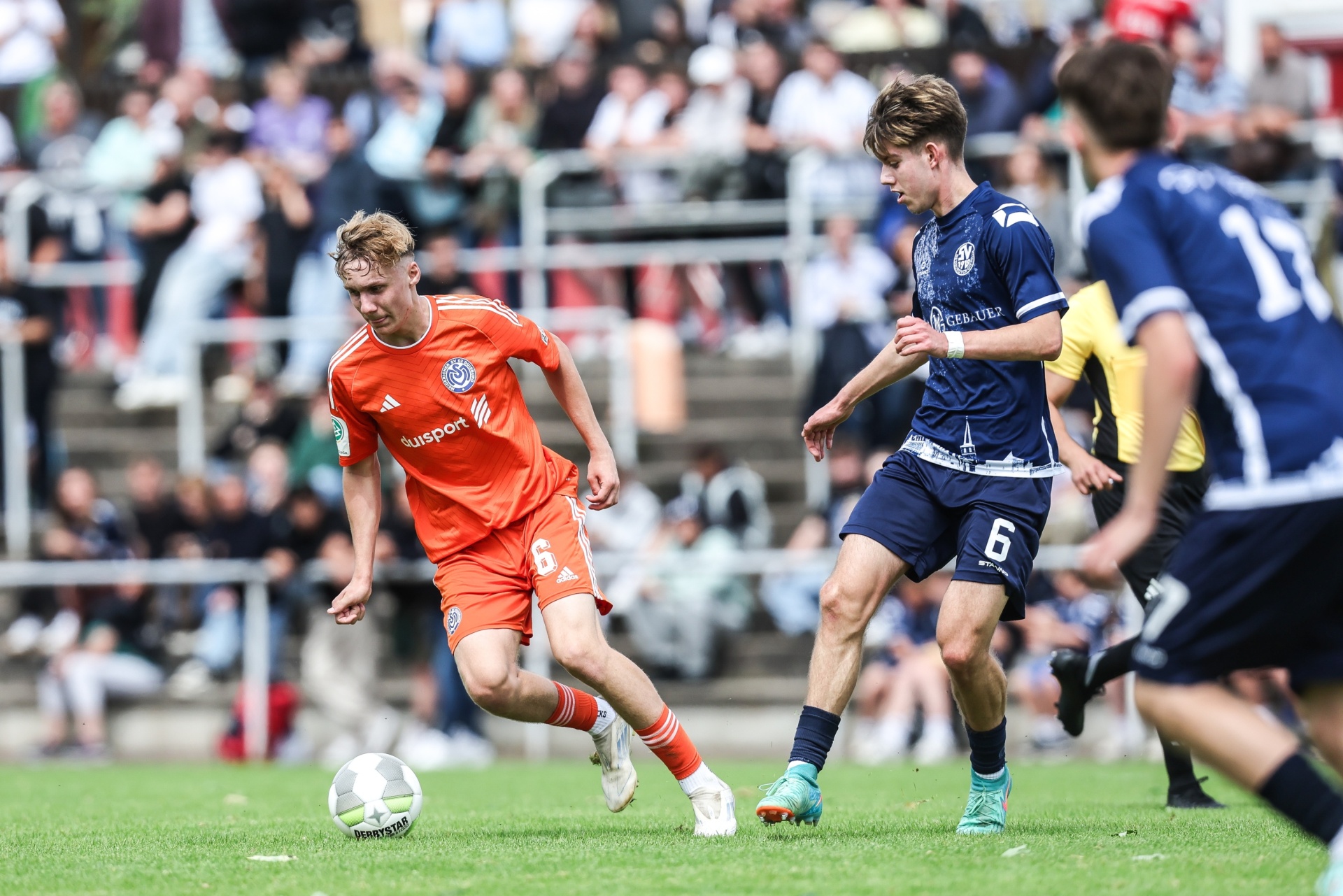 [{"clang_id":1,"value":"Fußballspiel: Zwei Spieler in orange und blau kämpfen um den Ball auf grünem Rasen. Im Hintergrund Zuschauer auf einer Tribüne."}]