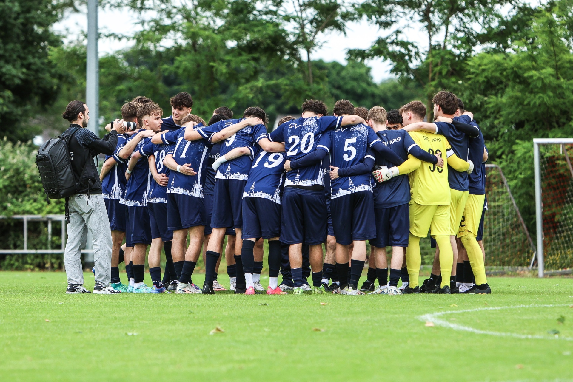 [{"clang_id":1,"value":"Fußballmannschaft in blau-weißen Trikots im Kreis, umringt von einem Trainer, auf einem grünen Fußballfeld. Im Hintergrund ein Fußballtor und Bäume."}]