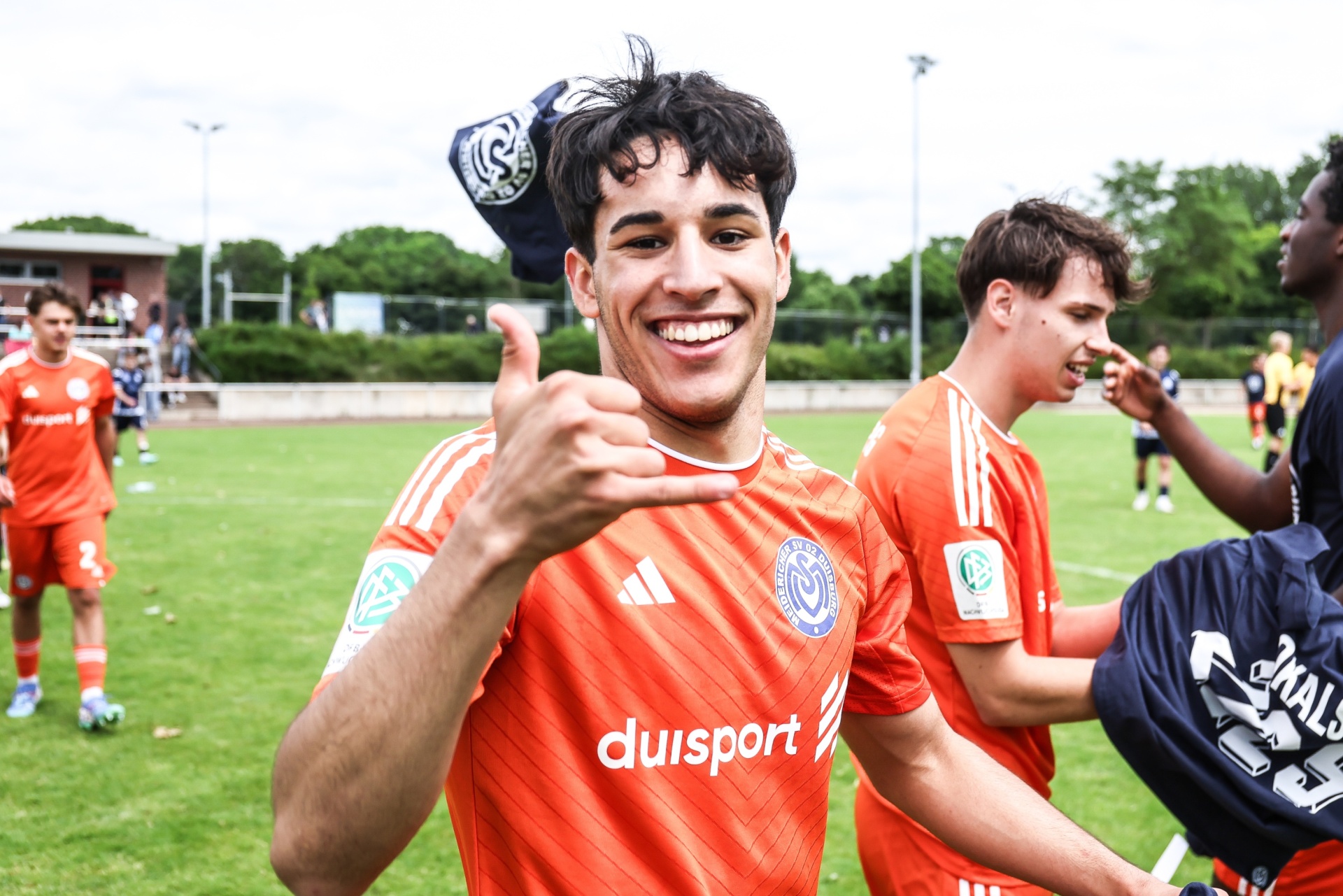 [{"clang_id":1,"value":"Junge Fußballspieler im orangefarbenen Trikot mit Sponsor-Logo \"duisport\". Vorderster Spieler lächelt und zeigt Daumen hoch. Hintergrund: Fußballfeld und Zuschauer."}]