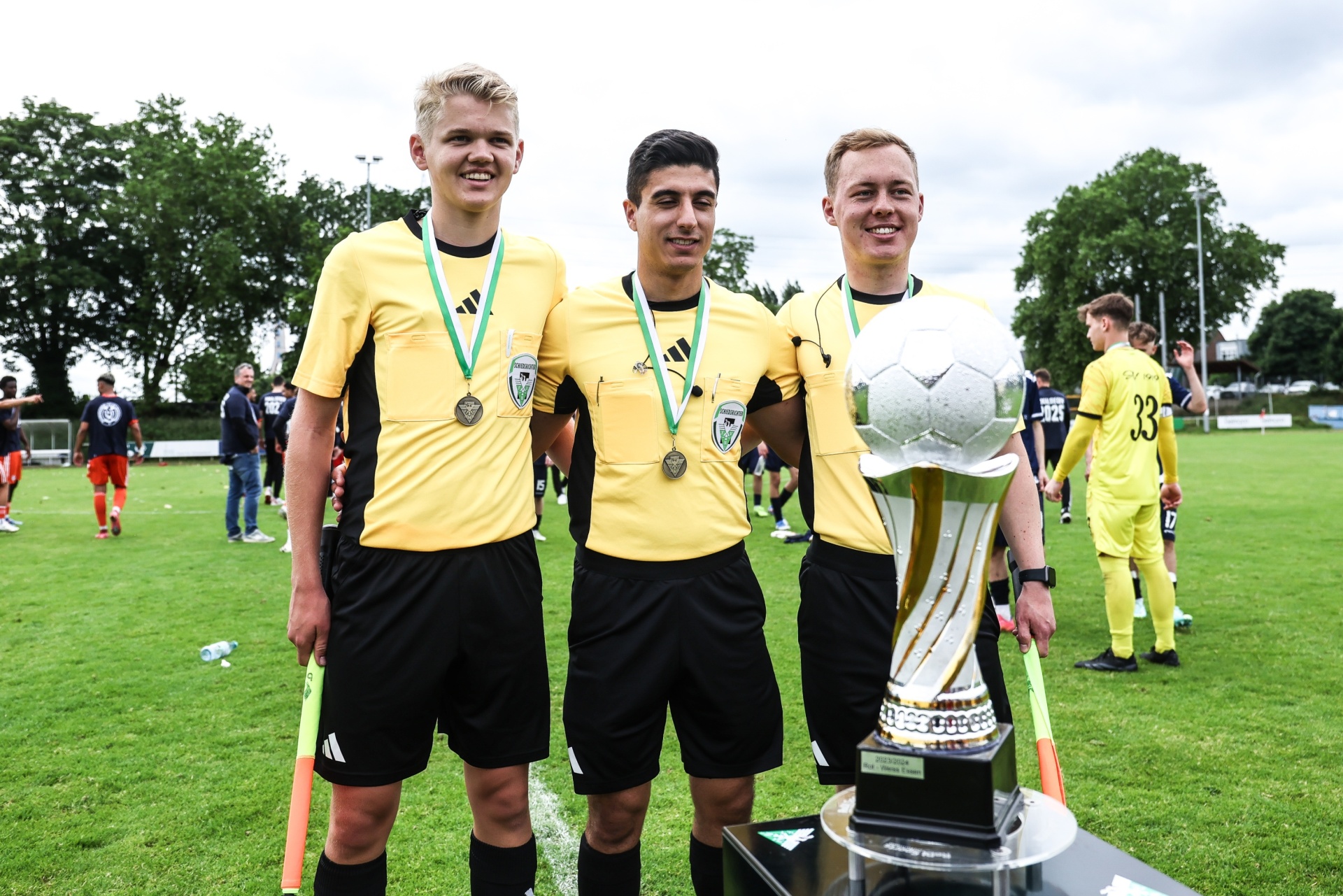 [{"clang_id":1,"value":"Drei Fußballspieler mit Medaillen und Trikots stehen vor einer silbernen Siegestrophäe auf einem Fußballfeld. Im Hintergrund sind weitere Spieler und Zuschauer zu sehen."}]