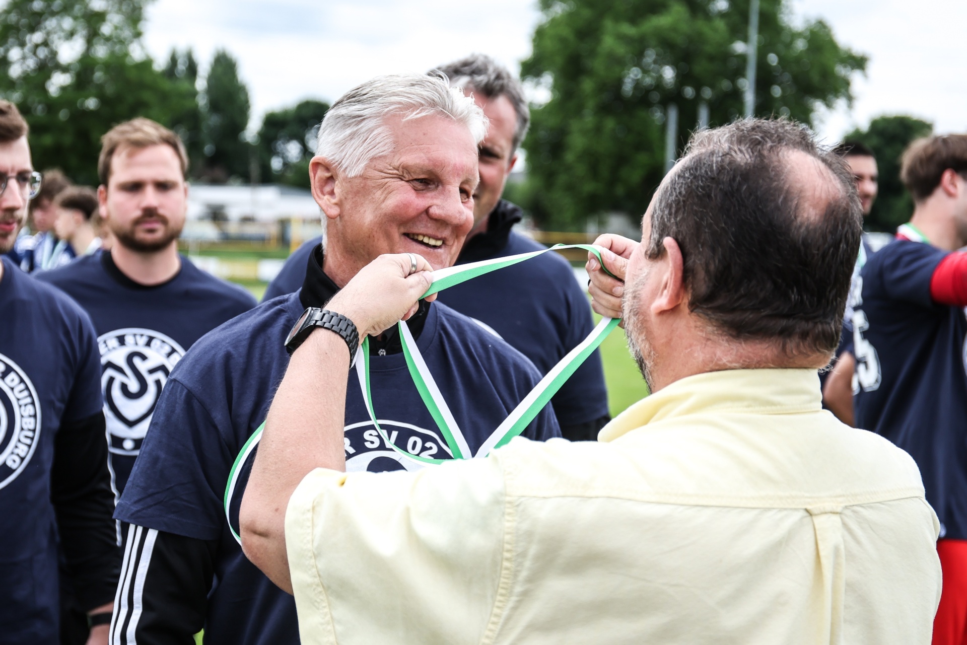 [{"clang_id":1,"value":"Zwei Männer bei einer Siegerehrung im Freien. Ein Mann in Sportkleidung erhält von einem anderen eine grüne Schleife um den Hals. Im Hintergrund weitere Personen und ein Fußballfeld."}]