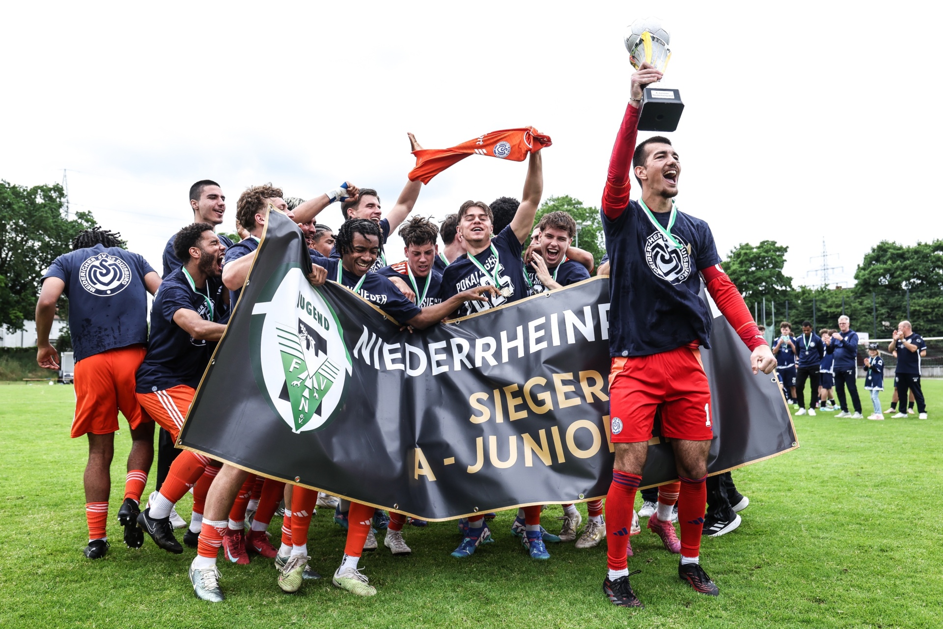 [{"clang_id":1,"value":"Fußballmannschaft feiert Sieg. Spieler mit rot-schwarzen Trikots und Flagge \"Niederrhein Sieger A-Junior\". Ein Spieler hält Pokal hoch."}]