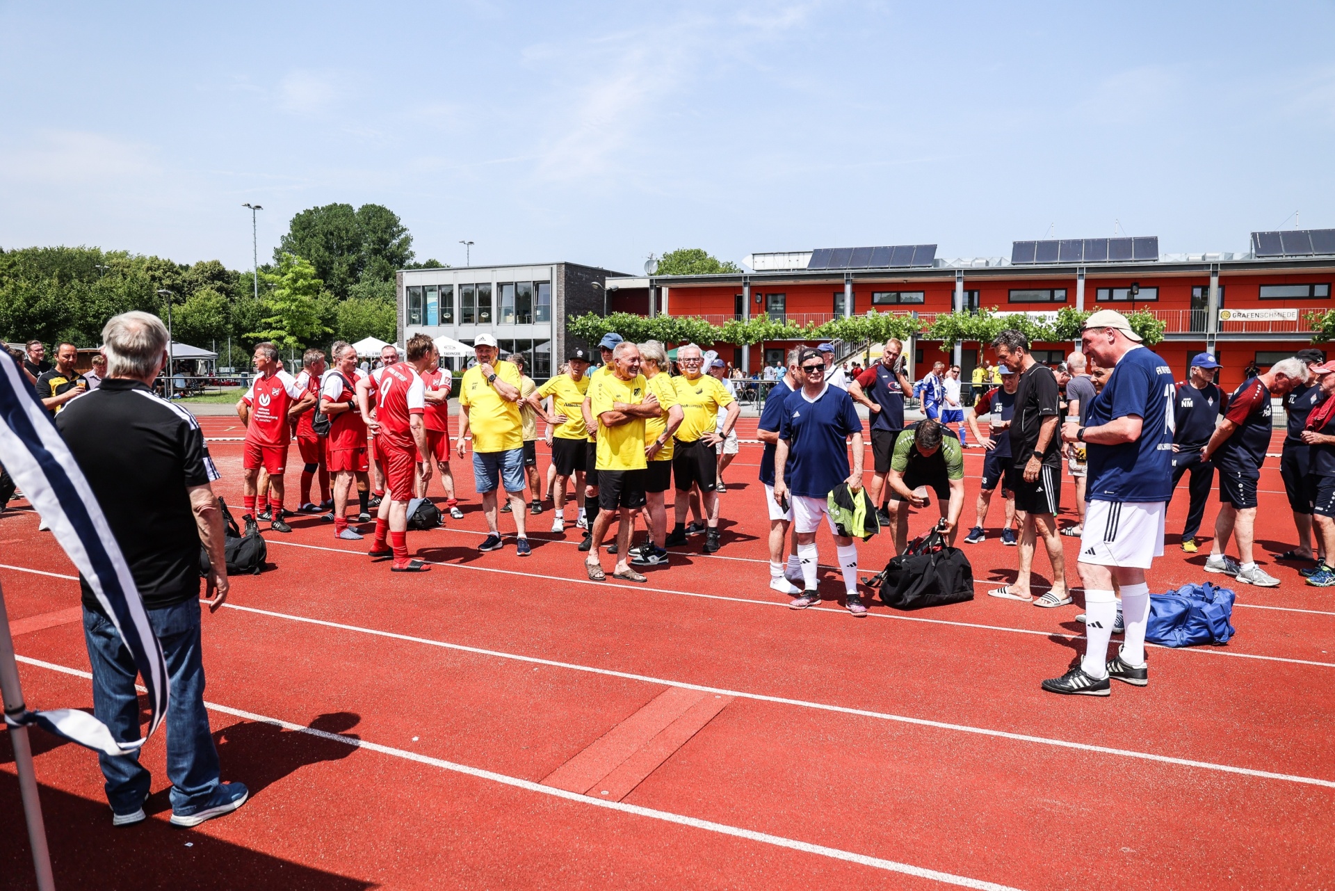 [{"clang_id":1,"value":"Eine Gruppe Sportler steht auf einer roten Tartanbahn vor einem Sportgebäude. Einige tragen gelbe T-Shirts, andere rote oder dunkle Kleidung. Ein Mann mit Kappe und Jacke steht ihnen gegenüber."}]