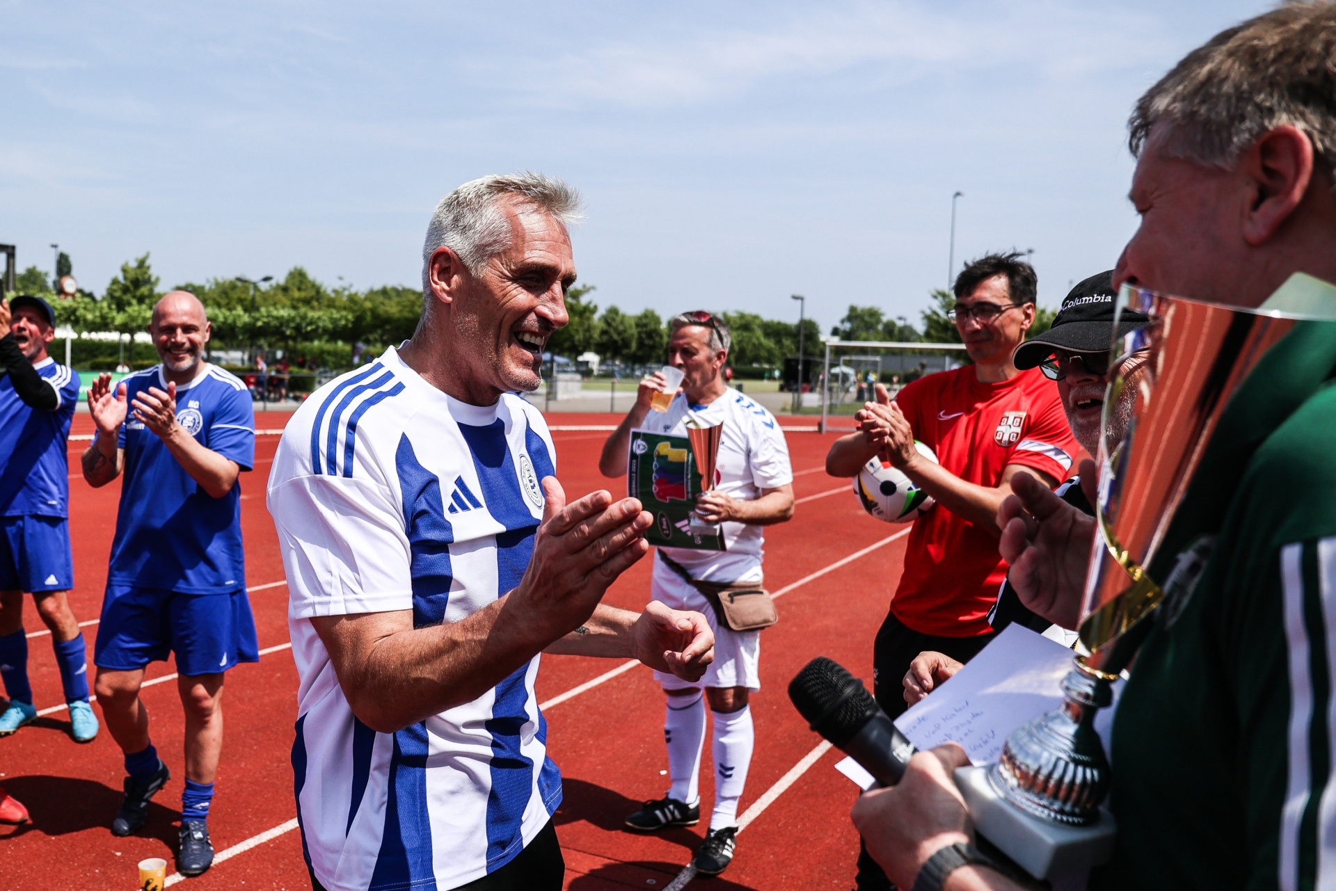[{"clang_id":1,"value":"Fußballfeld: Mann in blau-weiß gestreiftem Trikot wird interviewt, applaudiert. Im Hintergrund weitere Fußballspieler und Zuschauer."}]