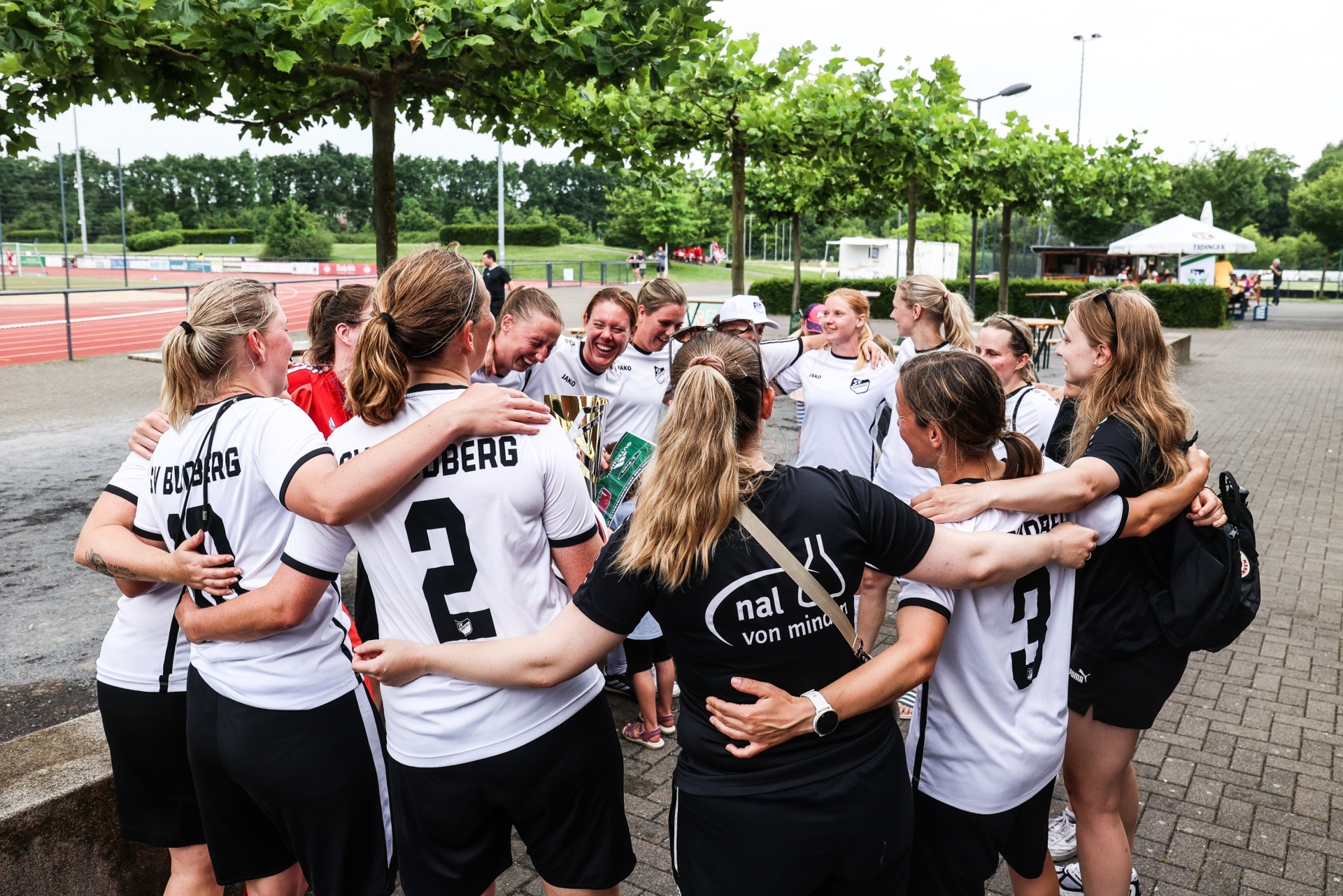 [{"clang_id":1,"value":"Fußballteam in schwarz-weißen Trikots feiert im Kreis, einige mit Pokal. Im Hintergrund Sportanlage und Bäume."}]